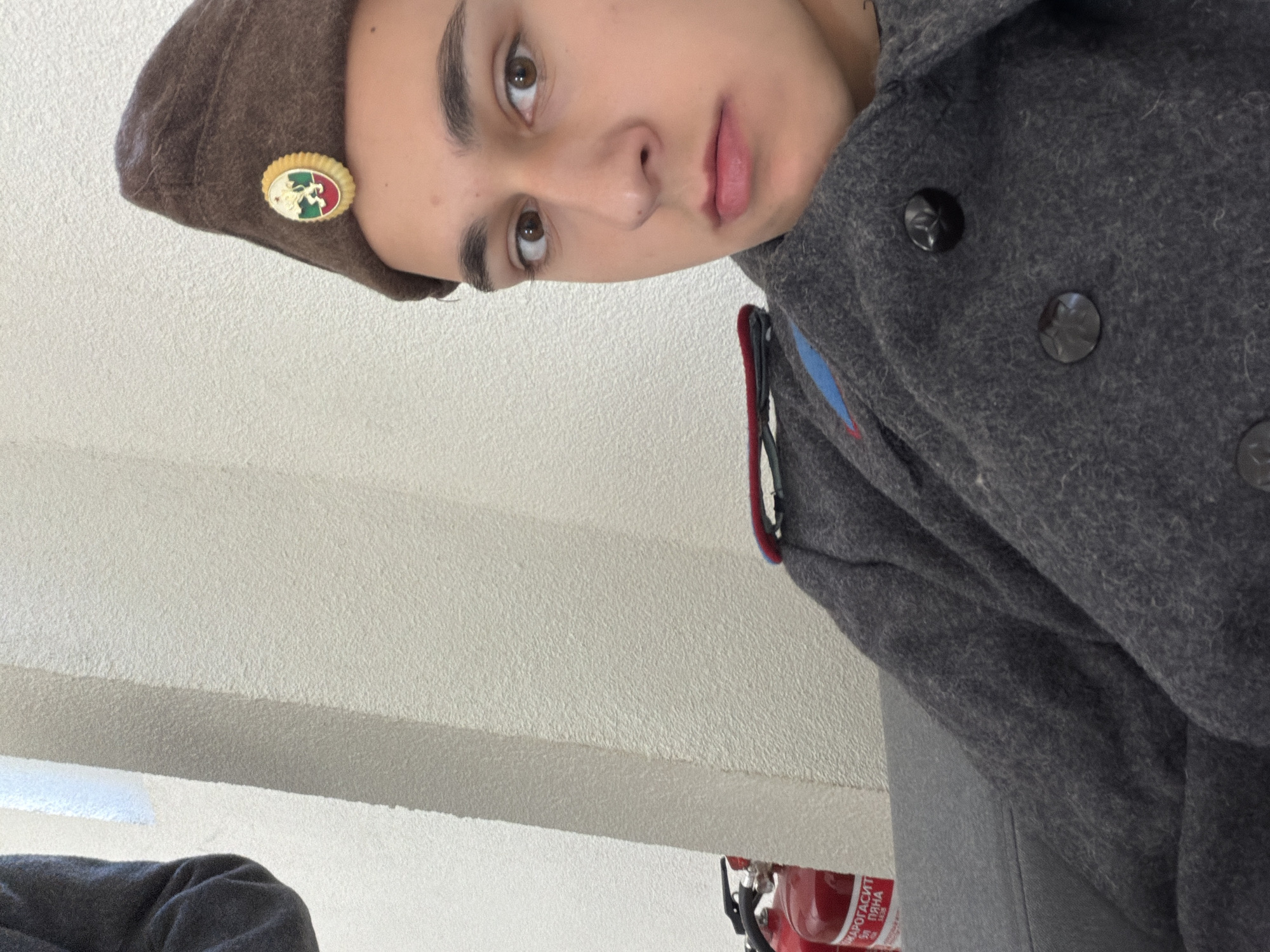 A person wearing a grey coat and a brown beret with a badge is visible in the image. The badge is oval-shaped with yellow trim and appears to have a white figure and what may be the colors of the Italian flag (green, white, and red). The coat has dark, round buttons and epaulets on the shoulders. Part of a red fire extinguisher is also visible. The location is Shumen, Bulgaria.