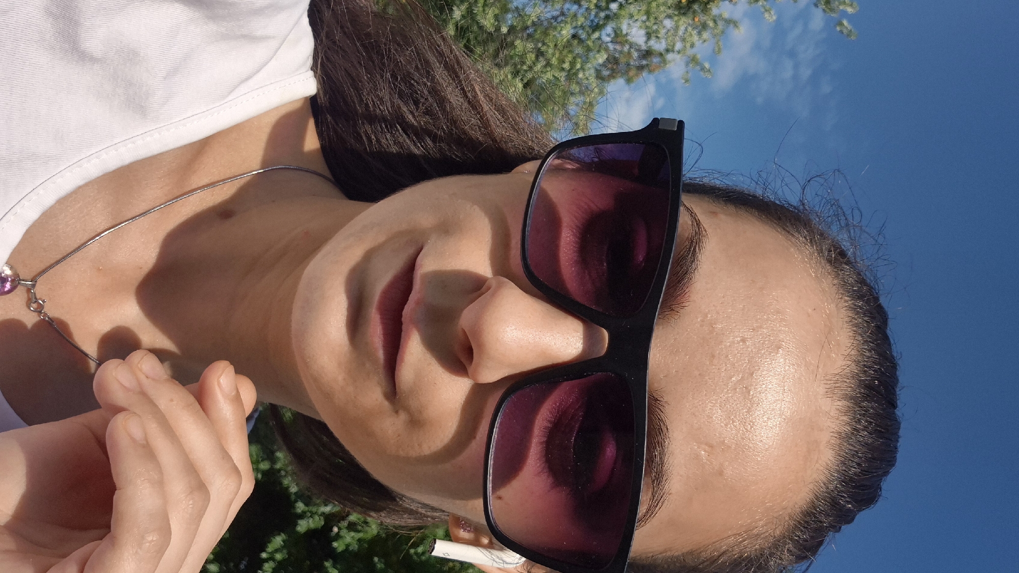 A medium shot photo shows a woman outdoors on a sunny day. She is wearing large black-framed sunglasses with pink lenses and a white t-shirt. She has dark hair pulled back and a silver necklace with a pink pendant. She is also wearing white wireless earbuds. The background is a blue sky with some clouds and green trees. The scene appears to be in a park or outdoor area in Sofia, Bulgaria.