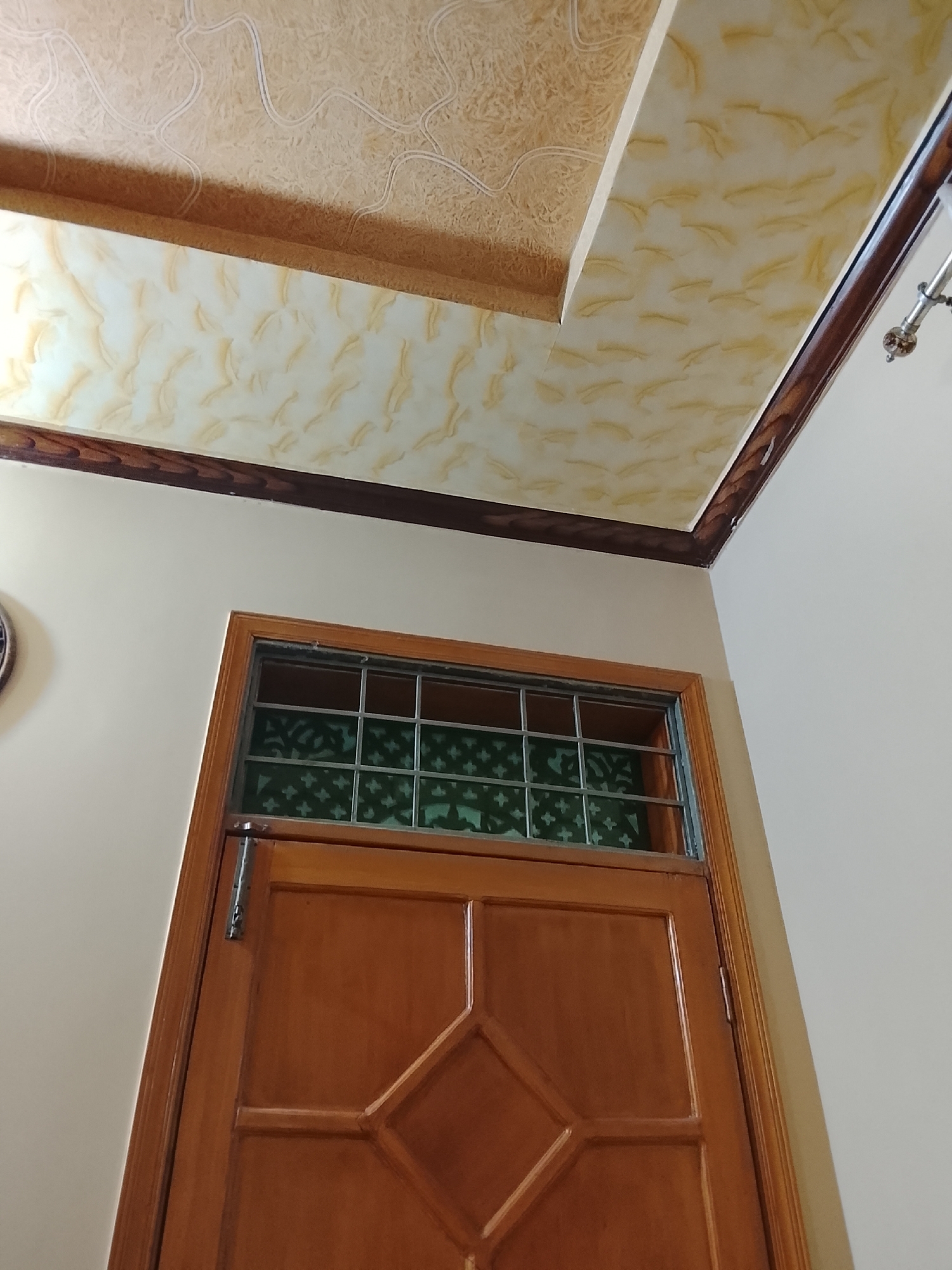 An indoor shot of a brown wooden door with a small barred window at the top, a latch on the left, and a geometric design in the lower half. The walls are painted a light beige or cream color. The ceiling has a patterned texture in shades of tan and yellow. The photograph was taken in a residential area in Multan, Pakistan.