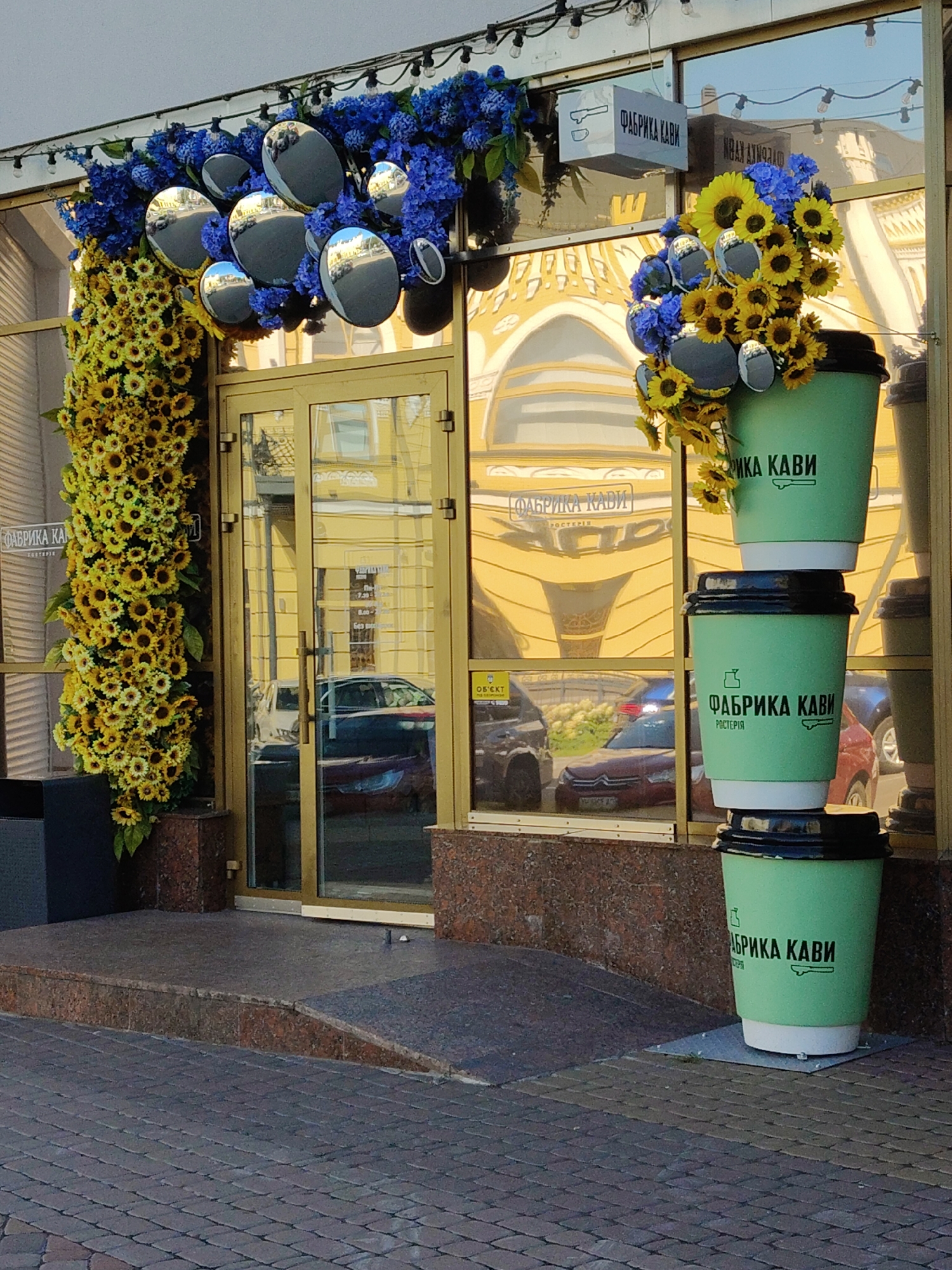 This is an eye-level, medium shot of the exterior of a cafe in Cherkasy, Ukraine, identified by signage as "Фабрика Кави" (Fabrika Kavi, meaning "Coffee Factory"). The cafe features a large glass storefront with a gold-colored frame. On the left, a tall, vertical arrangement of yellow sunflowers and blue flowers adorns the entrance. Above the door, a sign with a coffee cup and steam graphic is visible, along with the text "ФАБРИКА КАВИ". To the right of the entrance, three large, green disposable coffee cups, stacked vertically, serve as a decorative element. Each cup displays the "Фабрика Кави" logo and text. The cafe's facade is highly reflective, showing distorted images of the street and buildings opposite. The ground in front of the cafe is paved with brick-like stones, with a granite-covered ramp leading to the entrance. The overall scene suggests a vibrant and decorated commercial establishment.