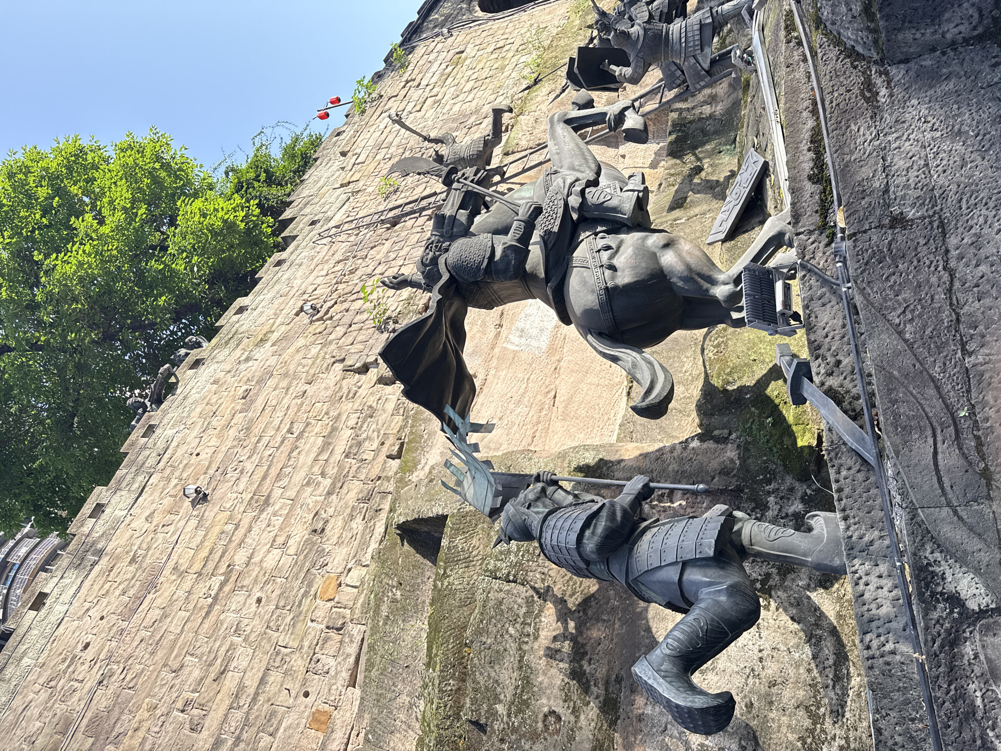 The image displays a dramatic upward-angled view of a large, textured stone wall adorned with numerous dark-colored statues of ancient warriors and cavalry. The scene is set outdoors under a bright, clear blue sky, indicating a sunny day.
The primary subjects are the dynamic statues, crafted from a dark material, possibly bronze or stone. A prominent equestrian statue depicts a warrior in full armor, wielding a spear, and mounted on a horse in mid-action, as if charging or ascending the steep wall. Below this, another armored warrior figure is seen, also in a dynamic pose against the rough stone, holding a weapon. Several other warrior figures and elements of battle, such as shields and banners, are integrated into the wall's design, suggesting a sprawling historical narrative or monument.
The wall itself is constructed from large, rough-hewn stone blocks, exhibiting various shades of brown and gray, with visible textures and some patches of moss or greenery. Green trees and dense foliage are visible along the top-left edge of the wall, contrasting with the ancient stonework. Two small, red, lantern-like objects hang from a structure near the top left, adding a cultural detail. The statues' intricate armor and weaponry, along with their dynamic poses, evoke a sense of strength, history, and movement. Given the location context of Yuzhong District, China, these statues likely depict figures or events from Chinese history or mythology. There is no visible text in the image.