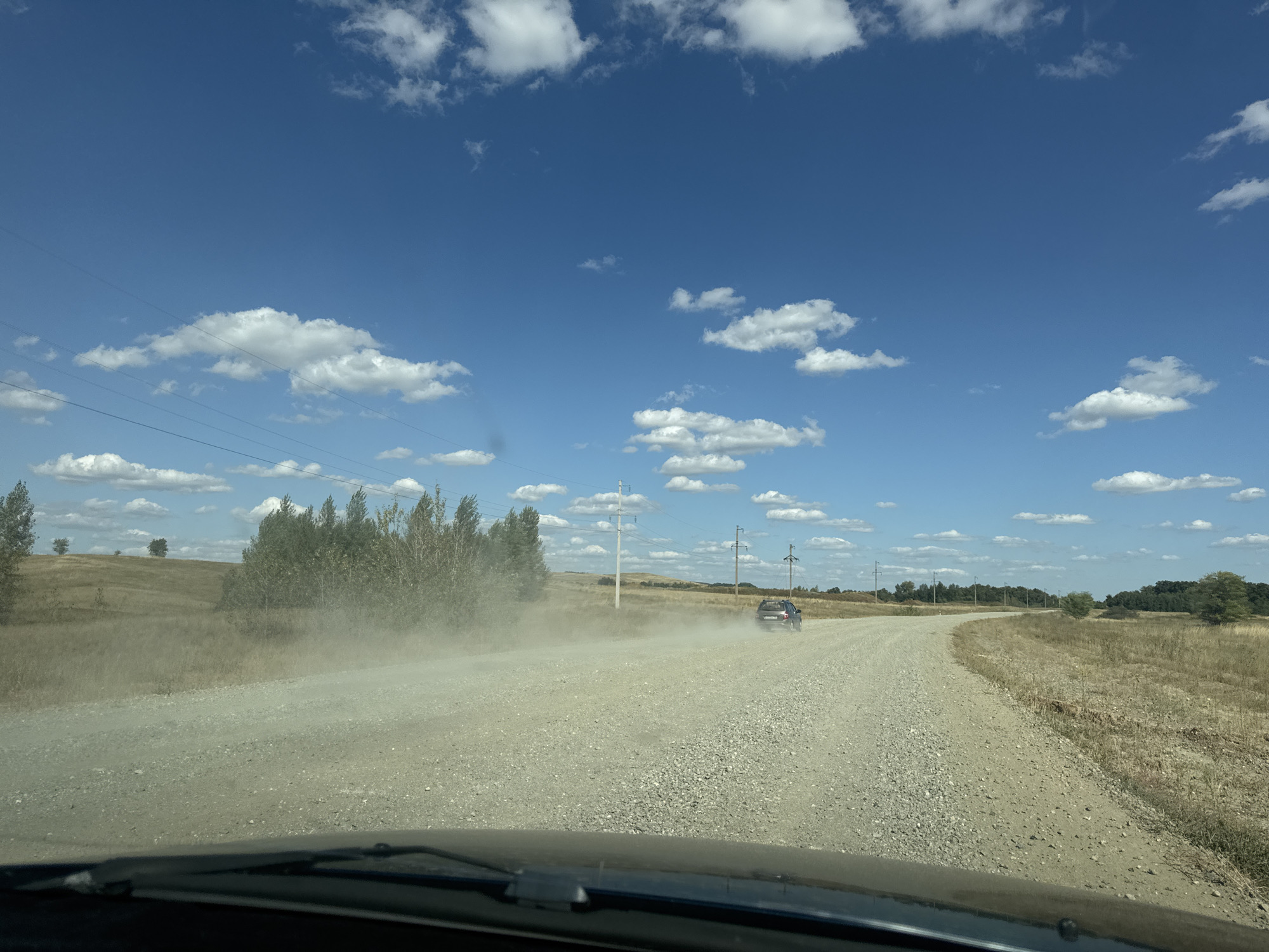 The image depicts a view from inside a vehicle, looking out onto a dusty, unpaved road in a rural setting. The road curves to the right, and a dark-colored SUV is visible driving away from the camera, kicking up a significant amount of dust. The landscape on either side of the road is dry and grassy, with scattered trees and shrubs. In the background, rolling hills are visible, and a line of utility poles runs along the side of the road. The sky is a bright blue with scattered white cumulus clouds. The scene is consistent with a summer day in a dry, agricultural region. The location is identified as Vasilievka, Russia, which suggests a rural area within that country. The presence of the car and the dust trail indicate recent activity. The overall impression is one of a journey through a sparsely populated, natural environment.