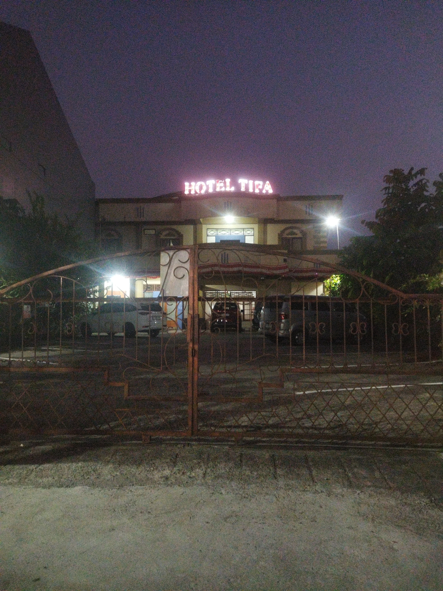The image depicts the exterior of Hotel Tifa in Wonogiri, Indonesia, at twilight. The hotel is identifiable by a large, illuminated sign displaying "HOTEL TIFA" in neon lights. The building features a pale facade with architectural details like arched windows and columns. A tall, dark structure, possibly another building or a large wall, is visible to the left.
In front of the hotel, a metal gate with decorative scrollwork is partially open, allowing a view of a parking area. Several vehicles, including a white SUV and other dark-colored cars, are parked within the premises. Some trees and foliage are present, with one large tree to the right of the building. The ground in the foreground appears to be a paved surface, possibly concrete or asphalt, with a rough texture. The overall lighting suggests it is either dusk or dawn, with artificial lights from the hotel and streetlights illuminating the scene against the dark sky. The image has a grainy quality.