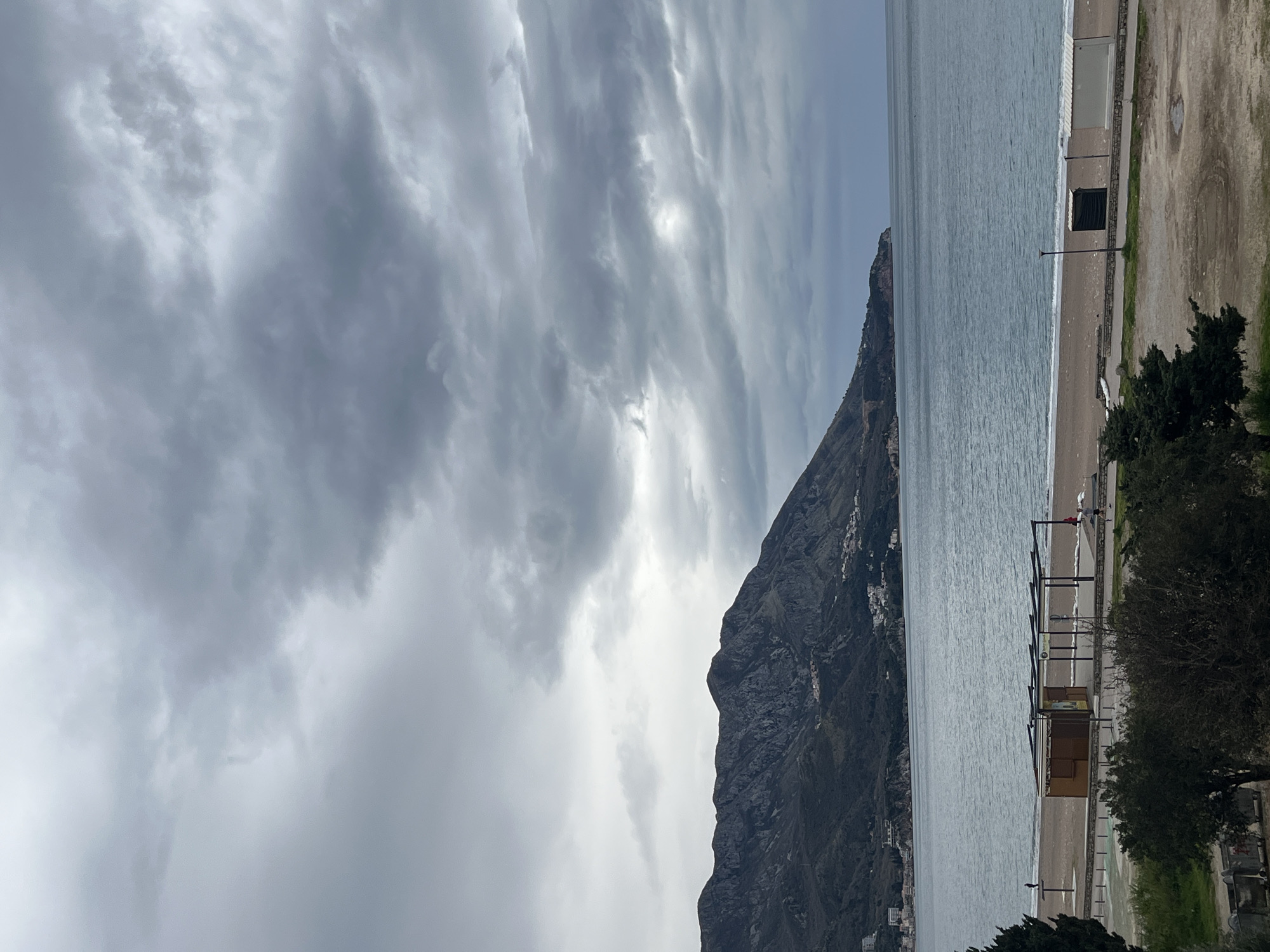 The image depicts a coastal scene in Boreti, Montenegro. A large, dark mountain rises from the sea, with some structures visible along its lower slopes. The sky is overcast with a mix of grey clouds. The sea is calm, with a sandy beach along the shoreline. Several wooden structures and trees line the beach. The adjacent land area is mostly bare ground with patches of grass. People are not clearly visible.