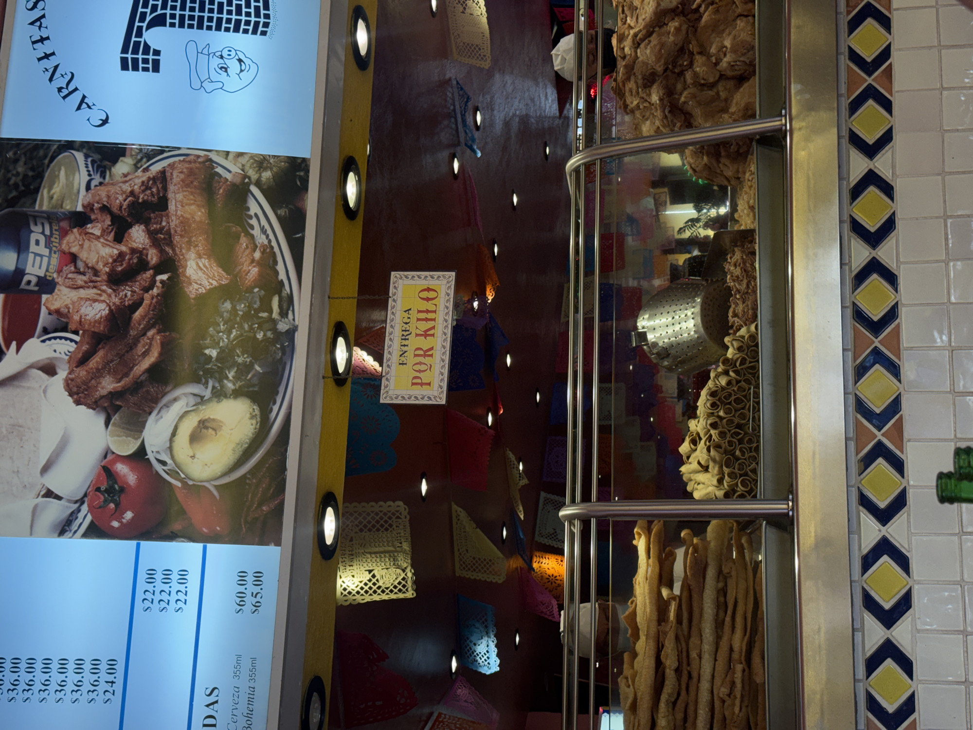 The image shows a food display at a restaurant in Puebla, Mexico. The menu on the left features "Carnitas," a traditional Mexican dish of braised or roasted pork, served with sides like avocado, tomatoes, and tortillas. Next to the menu is a sign indicating the price is "Por Kilo" (per kilogram). A display case features various fried foods, and the restaurant is decorated with strings of colorful, perforated paper banners, a common sight in Mexican establishments.
