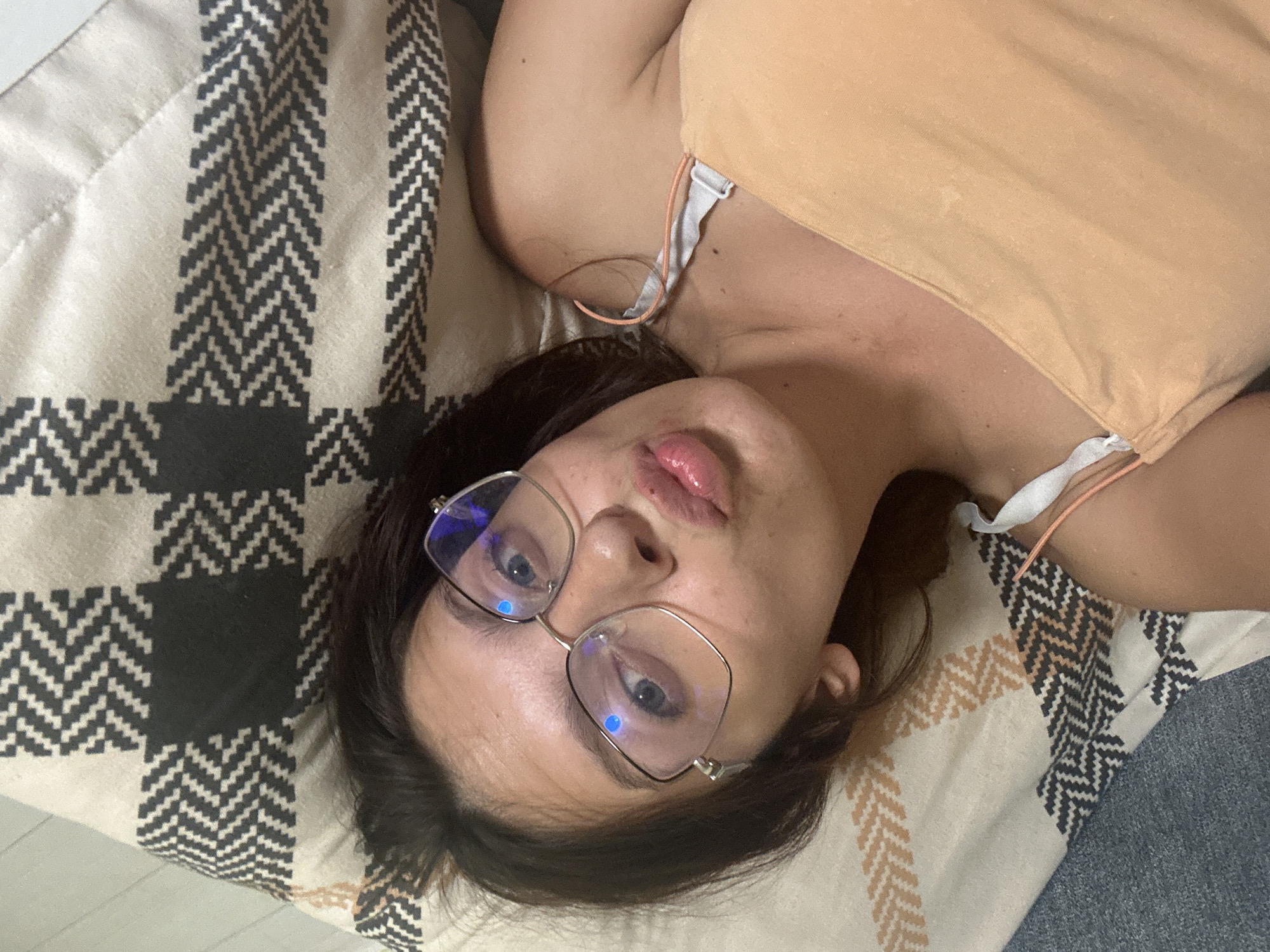 A woman with dark hair and blue eyes, wearing glasses and a peach-colored top, lies on a patterned blanket. Her lips are pursed as she looks at the camera. The blanket has a geometric design of black, white, and tan zigzags. The environment is indoors, with a portion of a blue surface visible at the bottom left. The scene does not provide any visual cues to determine its location within the city of Tryavna, Bulgaria.