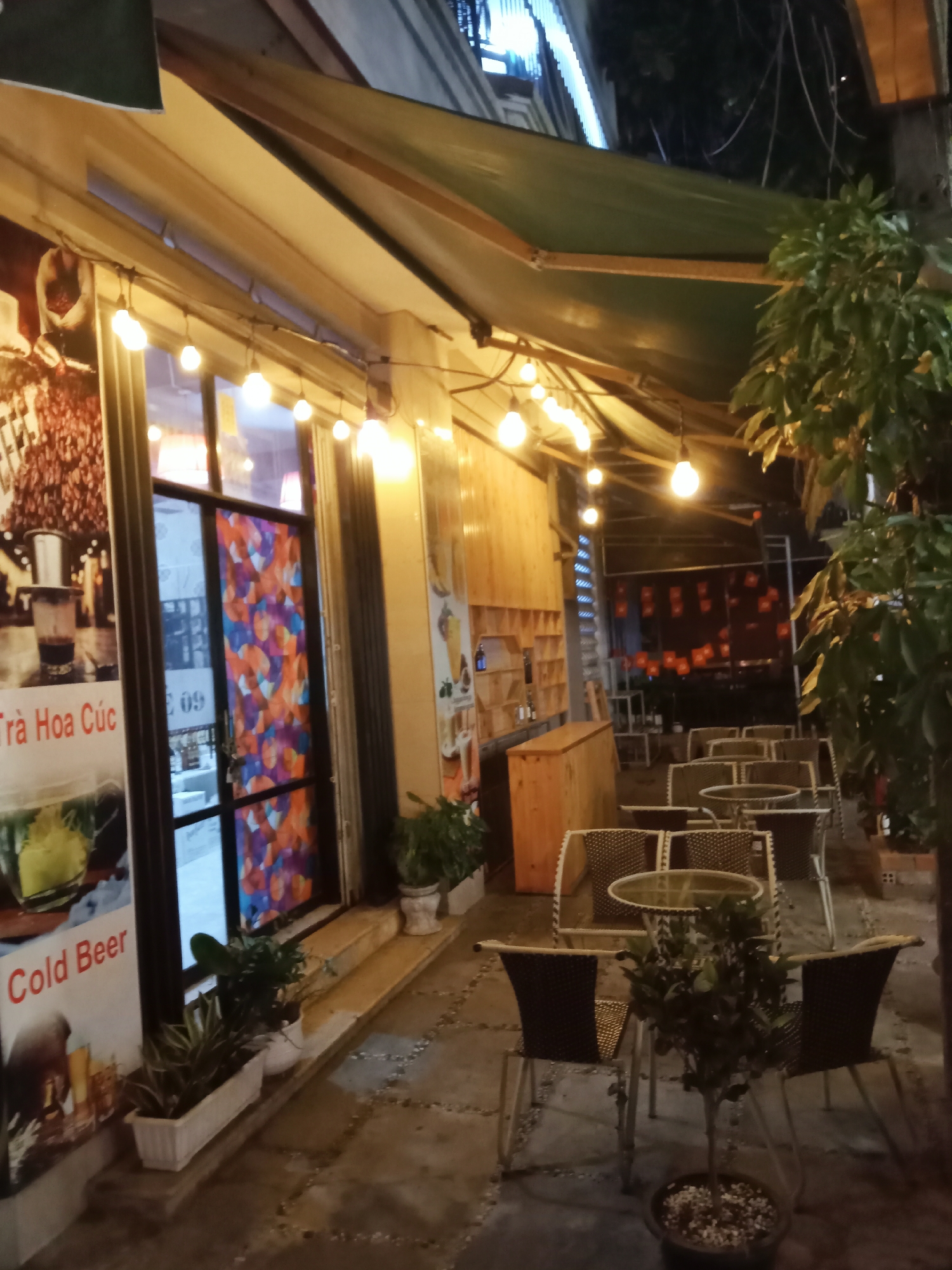 The image captures an outdoor seating area of what appears to be a cafe or restaurant at night. The scene is well-lit by numerous warm, glowing string lights draped overhead, creating a cozy and inviting atmosphere. A large green awning extends over the seating area, providing shelter.
The seating arrangement consists of several round tables with glass tops and matching wicker-style chairs, designed for outdoor use. There are no people visible in the image, suggesting the place might be closed or empty at the time of the photograph.
To the left, a building facade features large windows and a door with a colorful, abstract pattern. A prominent sign or poster next to the entrance displays text, including "Trà Hoa Cúc" (which translates to Chrysanthemum Tea) and "Cold Beer," indicating some of the offerings. Below the sign, a long planter filled with small plants is visible, and other potted plants are scattered around the seating area, including a small tree in a pot in the foreground.
Further inside the establishment, a wooden counter or bar area is visible, with shelves behind it. In the background, beyond the main seating, more tables and chairs are discernible, and a wall is decorated with multiple small red banners or flags, possibly Vietnamese flags given the context of Nha Trang. The ground is paved with patterned stones. The artificial lighting suggests it is evening or night time.