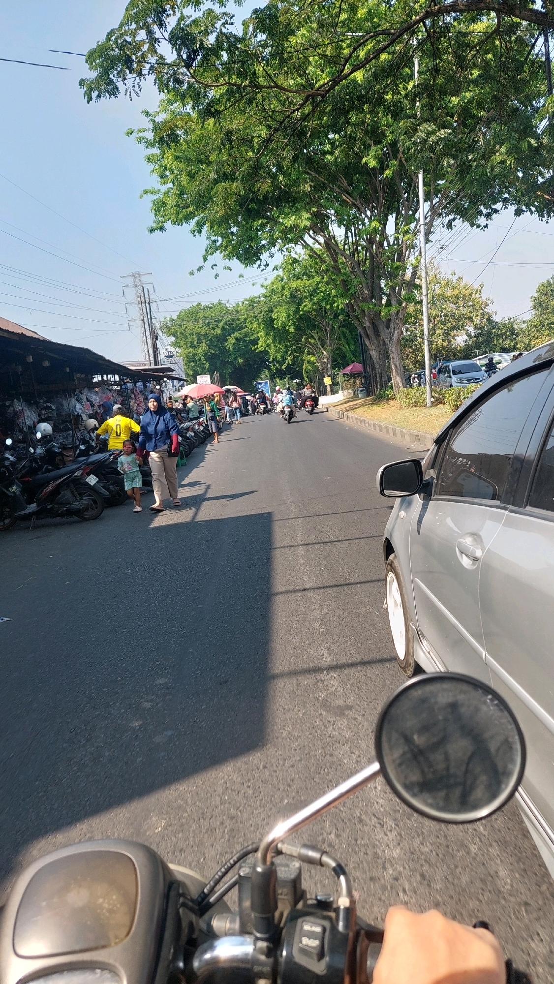 This image depicts a street scene in Sepande, Indonesia, on a sunny day. The perspective is from the viewpoint of someone riding a motorcycle, with the handlebars and a rearview mirror prominently in the foreground.
On the left side of the road, there is a row of market stalls with various goods displayed, including clothing and accessories. Several motorcycles are parked in front of the stalls. A woman wearing a blue hijab and light brown pants, accompanied by a young child in a light blue shirt and shorts, is walking away from the market stalls towards the street. Another person in a yellow shirt is standing near the market.
The road itself is paved and appears to be a two-lane street. Vehicles, primarily motorcycles and a silver car, are present on the road. The silver car is to the right of the frame, in the same lane as the motorcycle from which the photo is taken. Further down the street, more motorcycles and people are visible, some of whom appear to be riding or walking.
The environment is characterized by lush green trees lining the right side of the road, casting shadows on the asphalt. The sky is clear blue. Power lines and poles are visible in the background, indicating urban infrastructure. The overall impression is that of a bustling local market area in a residential or commercial district of Sepande.