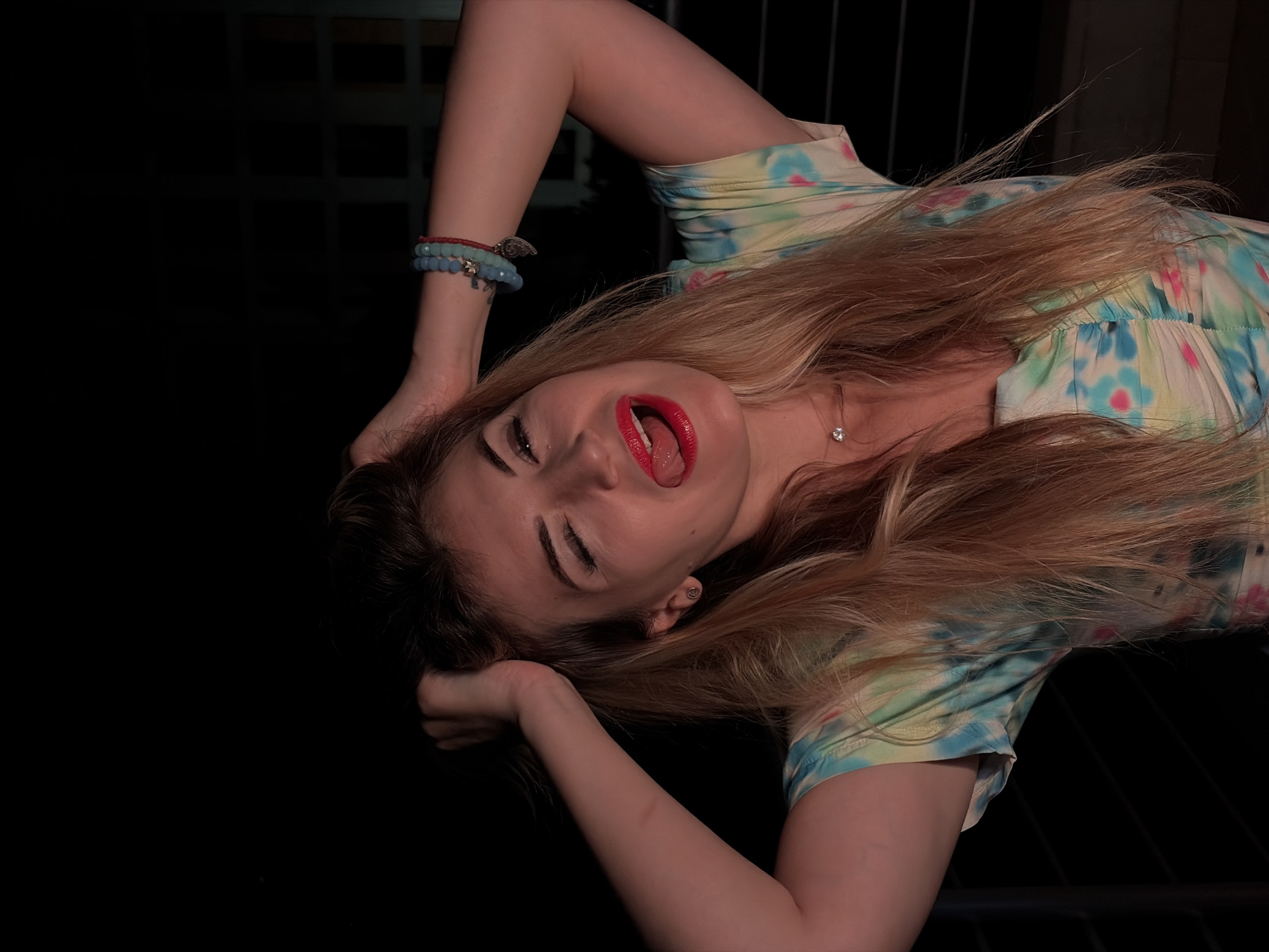 A woman with blonde hair and bright red lipstick is posing with her hands in her hair and her tongue sticking out, winking at the camera. She is wearing a tie-dye shirt and several bracelets on her wrist. The background is dark, with some hints of a building or structure visible. The image appears to have been taken at night, possibly on a balcony or terrace. The woman is positioned in the foreground, with the background out of focus. The image is framed to capture her face and upper body.
