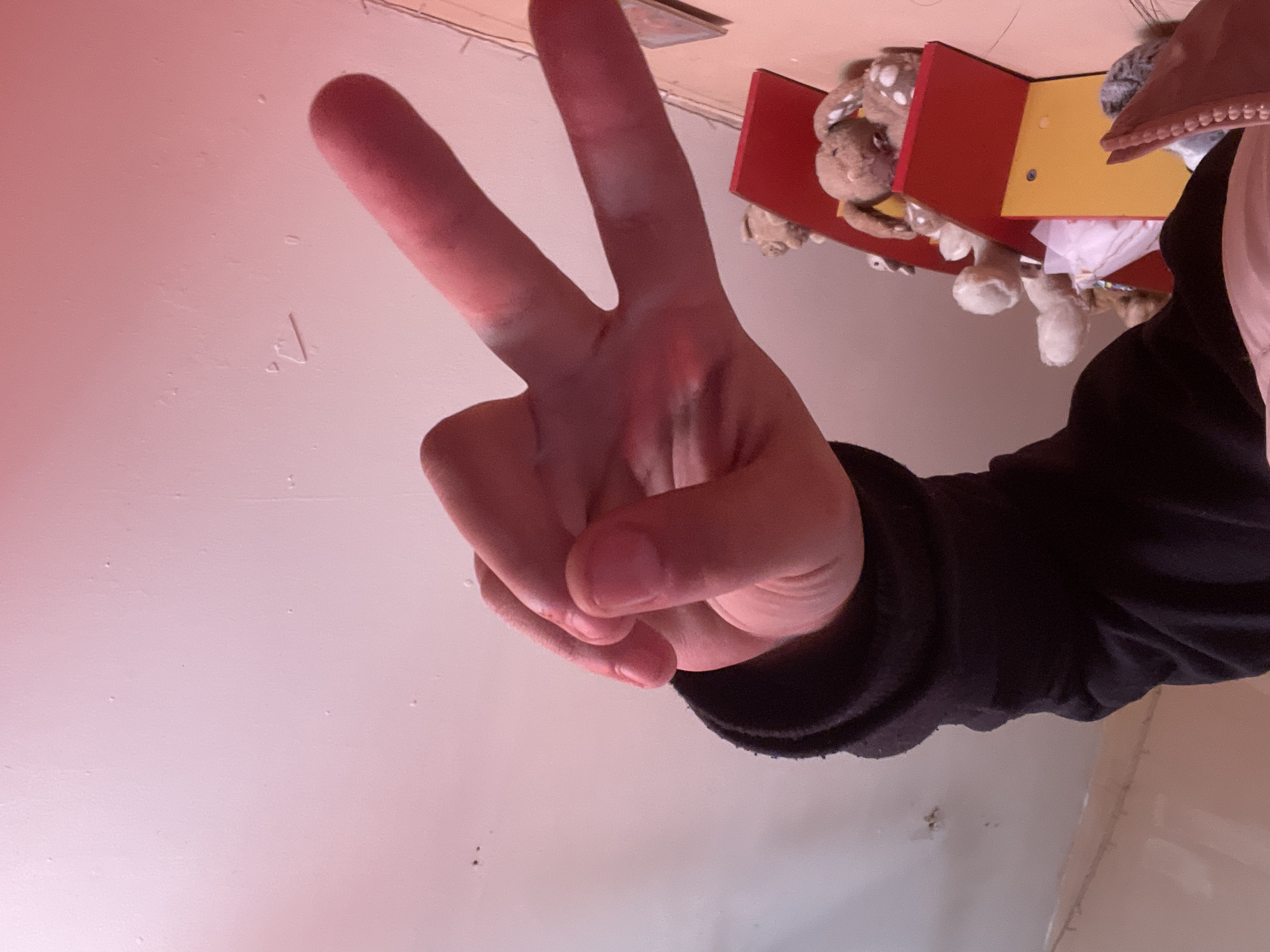 A hand is making the peace sign. The background includes a white ceiling, a red and yellow toy with plush toys, and a black sleeve. The location is Harmanli, Bulgaria.