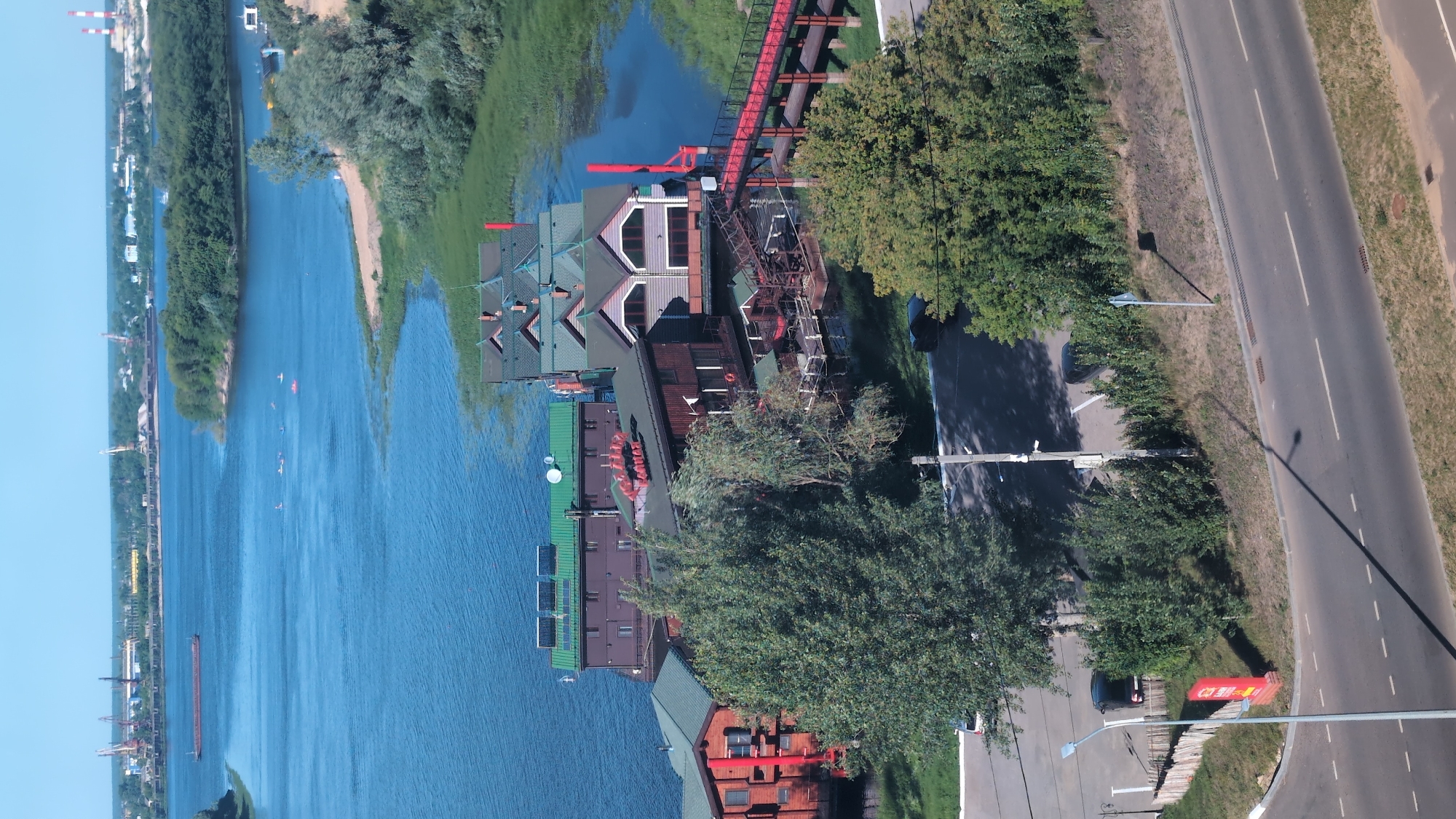 This is an aerial, daylight, wide-angle shot of a riverfront with several docked structures, likely a restaurant or hotel complex, on the water. The structures have multiple levels and red-tiled roofs. Some have solar panels and satellite dishes on their roofs. A red pedestrian bridge extends from one of the structures to the shore. The river is wide and blue, with a barge visible in the distance and several small boats with people in them closer to the shore. The riverbank is lined with trees and vegetation. In the foreground, a road with a few parked cars and a single streetlight is visible. The city of Nizhny Novgorod, Russia, is in the background, with industrial cranes and chimneys visible on the horizon. The scene appears to be a recreational area on the Volga River.