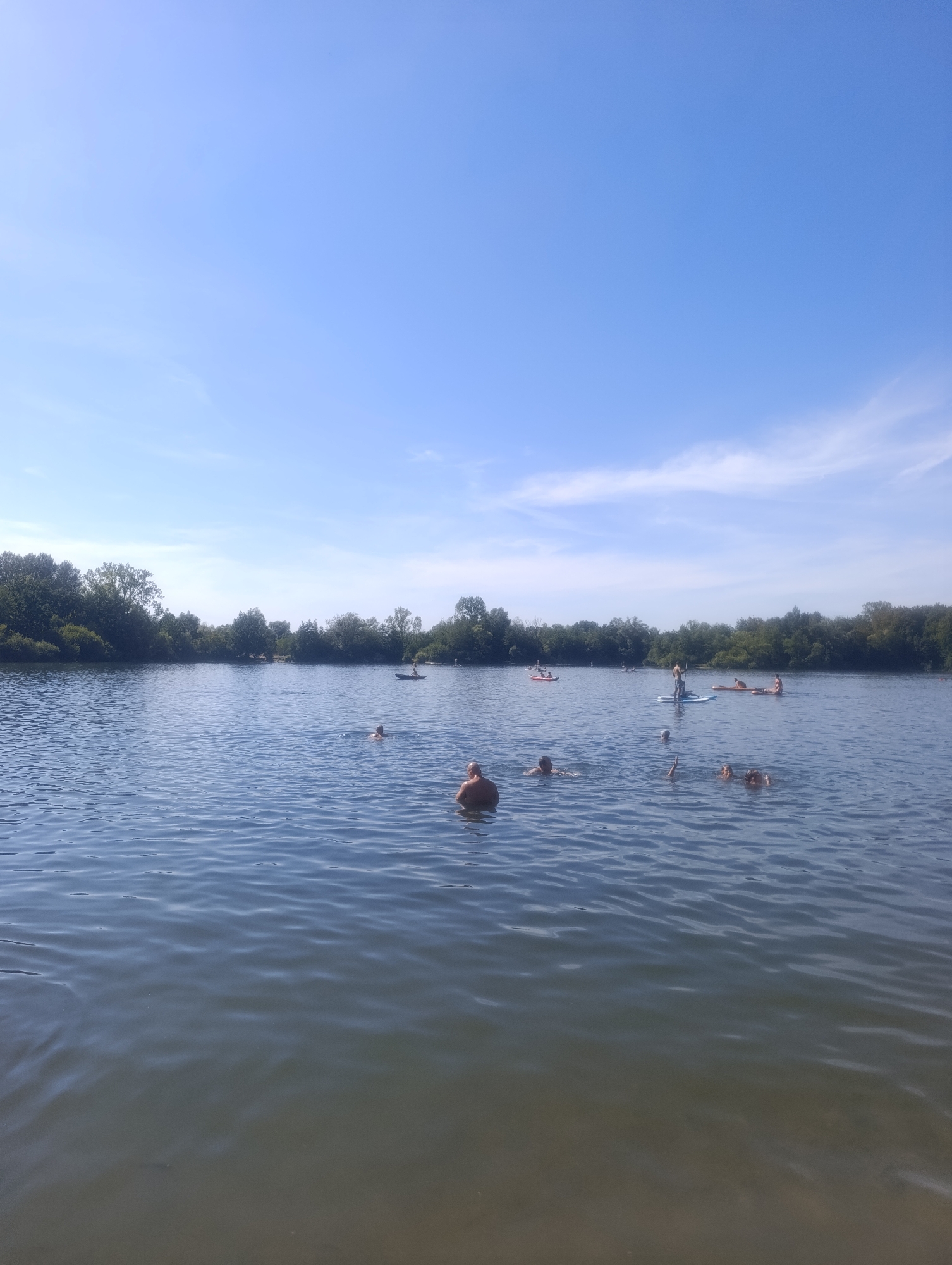 The image depicts a sunny day at a body of water, likely a lake, with several people engaged in recreational activities. In the foreground, two individuals are partially submerged in the water, with one person appearing to be facing away from the camera and the other to their side. Further out on the water, a person is standing on a paddleboard. In the middle ground, several other individuals are visible, some swimming and others on paddleboards. In the background, a dense line of trees forms the shoreline. The sky is clear blue with a few wispy white clouds. The location is identified as Křelov, Czechia.