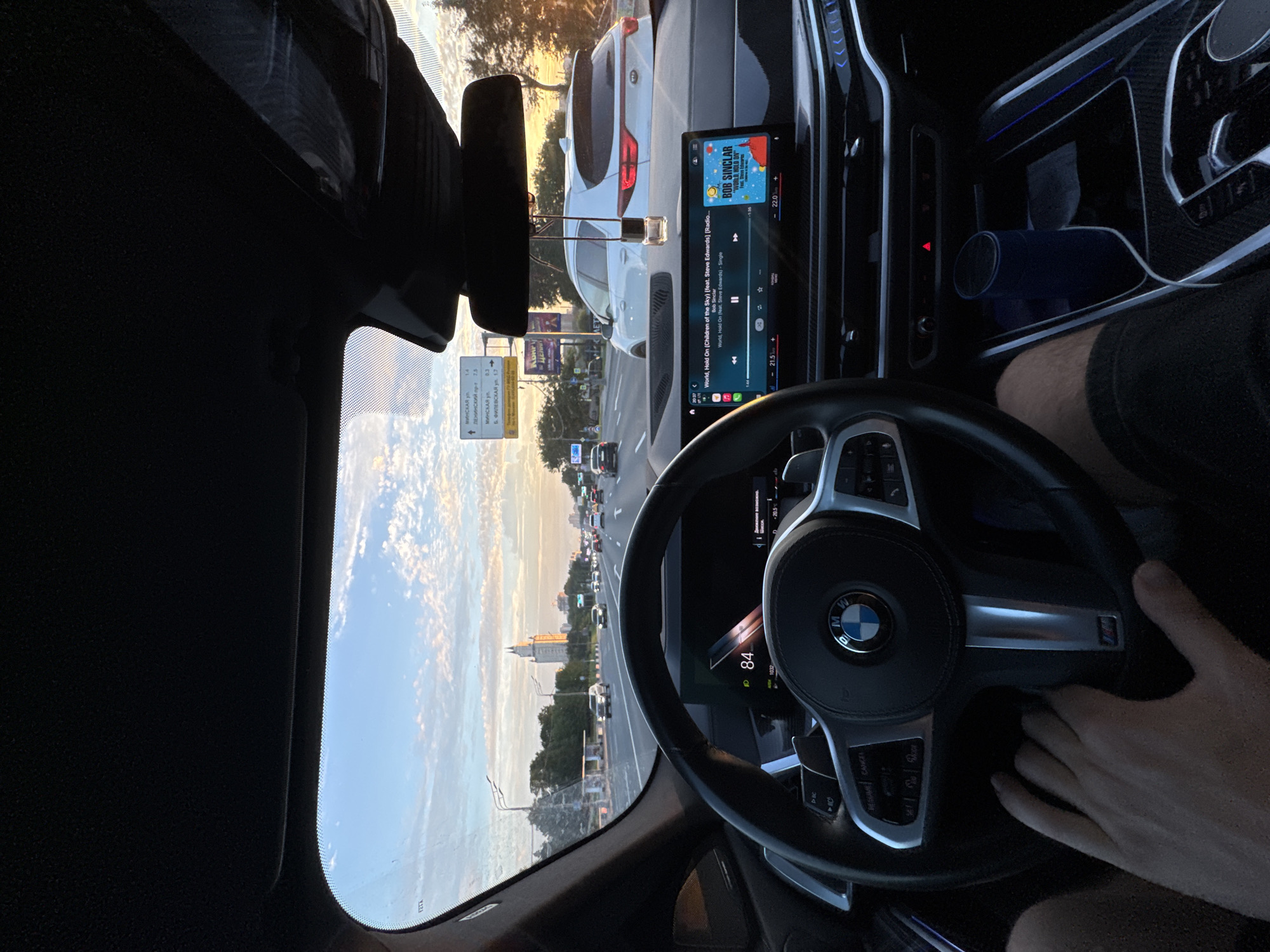 The image is taken from the perspective of a driver in a BMW vehicle, looking forward through the windshield and at the dashboard. The vehicle is on a multi-lane road, with traffic visible in front and to the sides. The sky is partly cloudy with a sunset glow. A tall, distinctive building with a spire is visible in the distance. Road signs with Russian text, including "Leninskiy pr-t," are present. The car's infotainment system displays song information for "World, Hold On (Children of the Sky)" by Bob Sinclar. The driver's hand is visible on the steering wheel. The scene appears to be in an urban or suburban area.