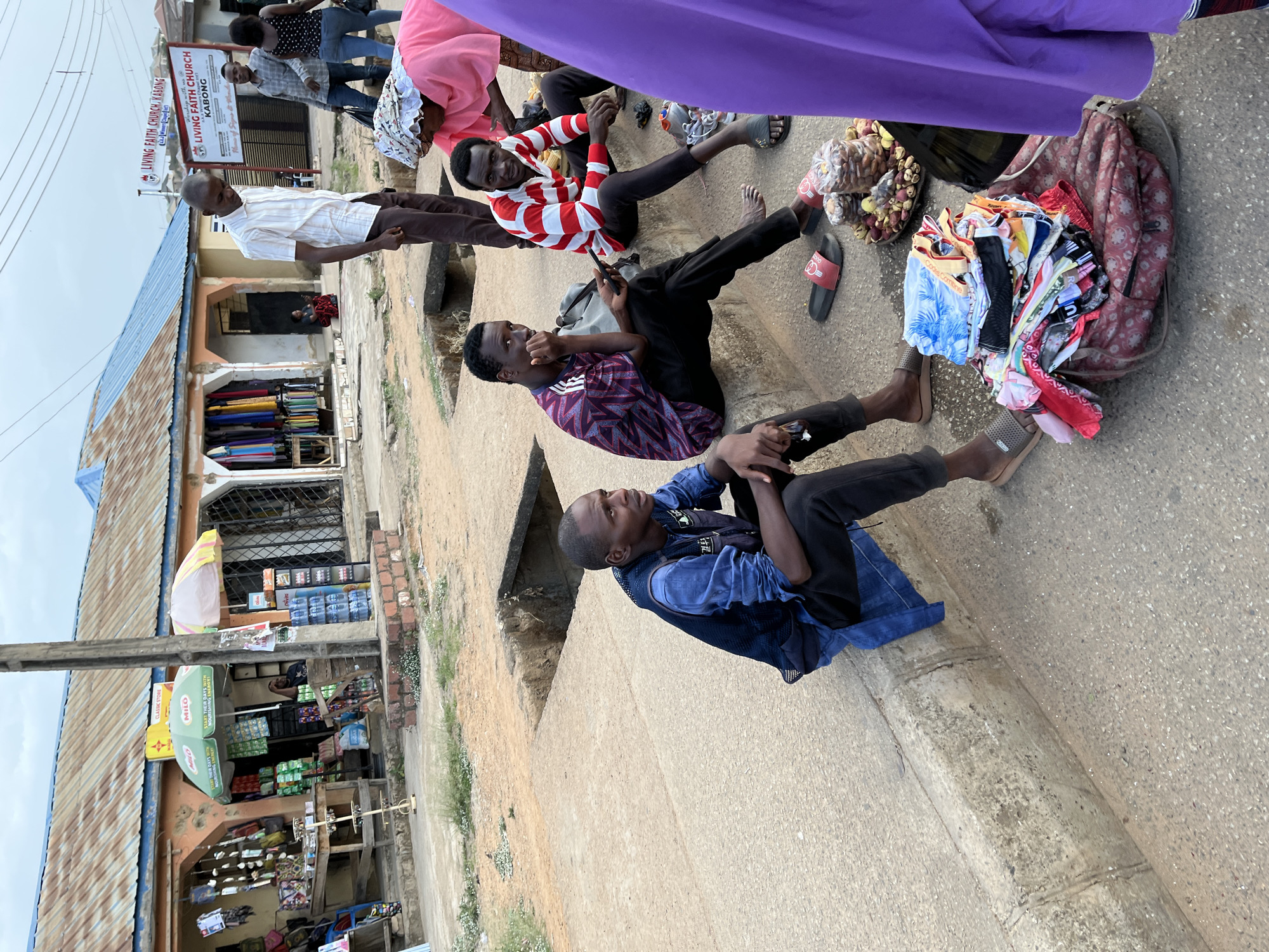 The image depicts a street scene in Jos, Nigeria. Three young men are seated at the edge of a concrete structure, likely a sidewalk or raised platform, in an informal gathering. The man closest to the camera wears a denim jacket and black pants, while the second man has on a maroon and navy patterned shirt and black pants. The third man is clad in a red and white striped shirt with black pants.
Adjacent to the men are goods for sale, including what appears to be a pile of clothes and some nuts. A person draped in purple fabric partially obscures the upper portion of the image.
Behind the men, there's a row of shops and buildings. The storefronts display various items including clothes and groceries, with visible signs like "CLASSIC STORE" and "MILO." A sign for "LIVING FAITH CHURCH, KABONG" is visible above one of the buildings. The sky is overcast, suggesting it may be a cloudy day.