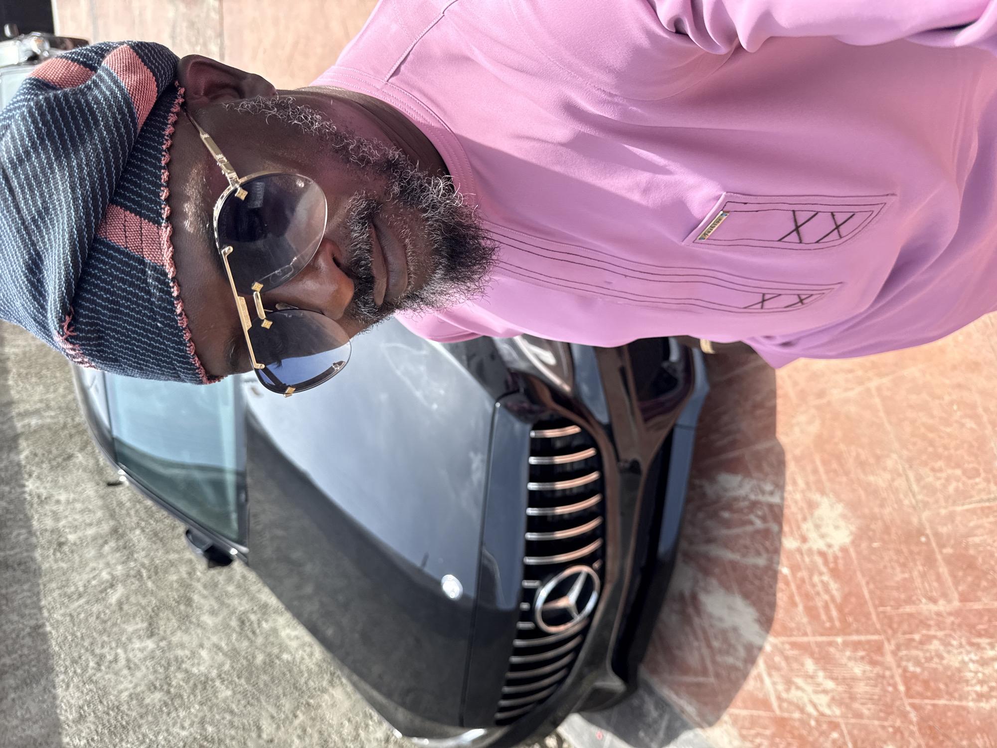 A man wearing a patterned cap, sunglasses, and a pink shirt stands next to a black Mercedes-Benz. The scene is located in Igboefon, Nigeria, as context was included in the instructions. The ground appears to be tiled.