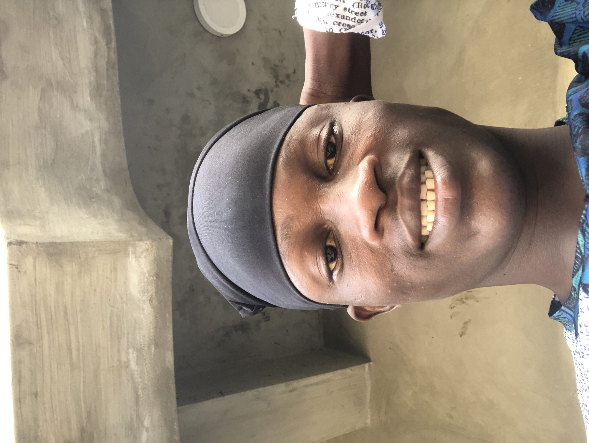 The image is a selfie taken outdoors. The subject is a young Black male, facing the camera, with a black durag on his head and a blue and green patterned shirt. He is smiling, revealing his teeth. His right arm is visible, resting on his head, with a white cloth with black text wrapped around his wrist. The background appears to be an outdoor wall and ceiling structure made of concrete. The lighting suggests it is daytime. The location is Ado, Nigeria.