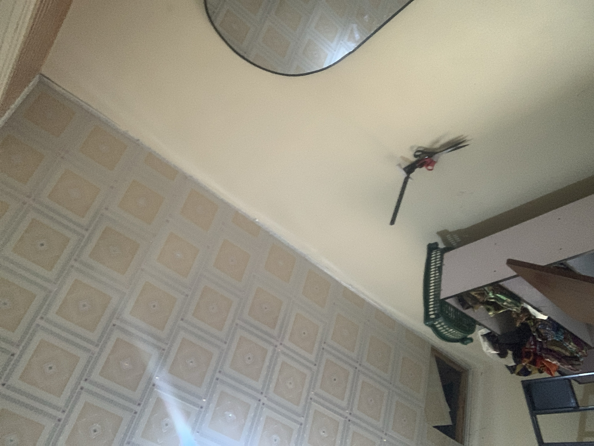 The image depicts an indoor scene, likely a room, with a patterned ceiling and a light-colored wall. In the lower half of the image, a tall piece of furniture, possibly a cabinet or wardrobe, is visible, laden with colorful fabrics and a green plastic basket. On the wall, a set of keys with a red fob is hanging. A large mirror with a black frame is also partially visible on the right side of the wall, reflecting the patterned ceiling. The overall impression is of a domestic interior, possibly cluttered. The location is identified as Jauro Yinu, Nigeria.