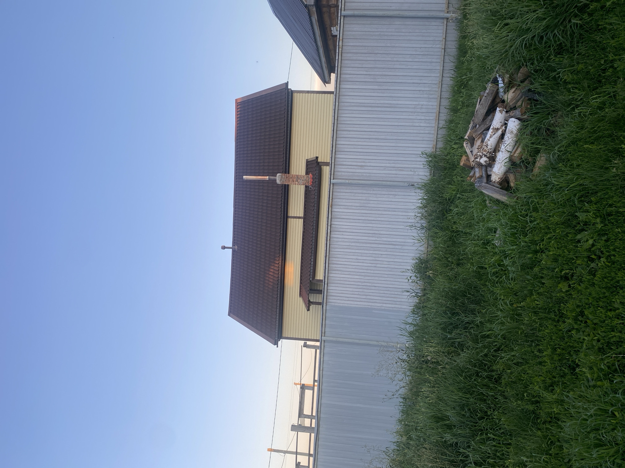 In the city of Verkhniy Karbush, Russia, an outdoor scene depicts a residential area. The image captures a clear, pale blue sky above two houses. The house in the foreground is a two-story structure with yellow siding and a dark brown, angled roof. A brick chimney with a metal pipe extends from the roof. To the right of this house, a portion of another house with a darker, angled roof and log construction is visible. A tall, corrugated metal fence runs horizontally across the midground, separating the houses from the foreground. The foreground is filled with tall, green grass and a pile of chopped wood, some of which appears to be painted white. The lighting suggests it is either early morning or late evening, with a warm glow on the roof of the yellow house. The overall environment appears to be rural or suburban.