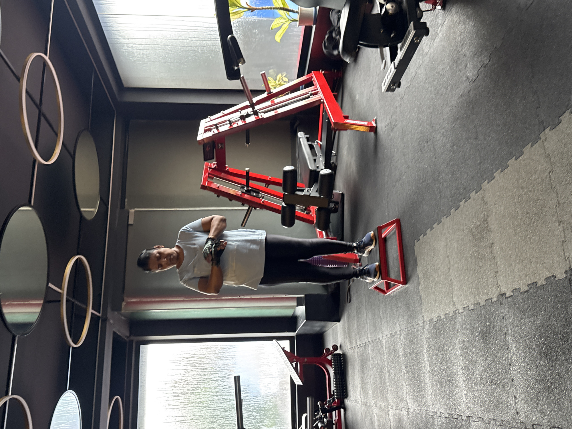 A person stands on a red exercise step in a gym. The person is wearing a light blue t-shirt, black leggings, black gloves, and athletic shoes. The gym has dark grey flooring made of interlocking mats, and a large window with textured glass behind the person. To the right, a red weight-lifting rack and other gym equipment are visible. Overhead, circular light fixtures are mounted on the dark ceiling. The scene is located in Kolkata, India.