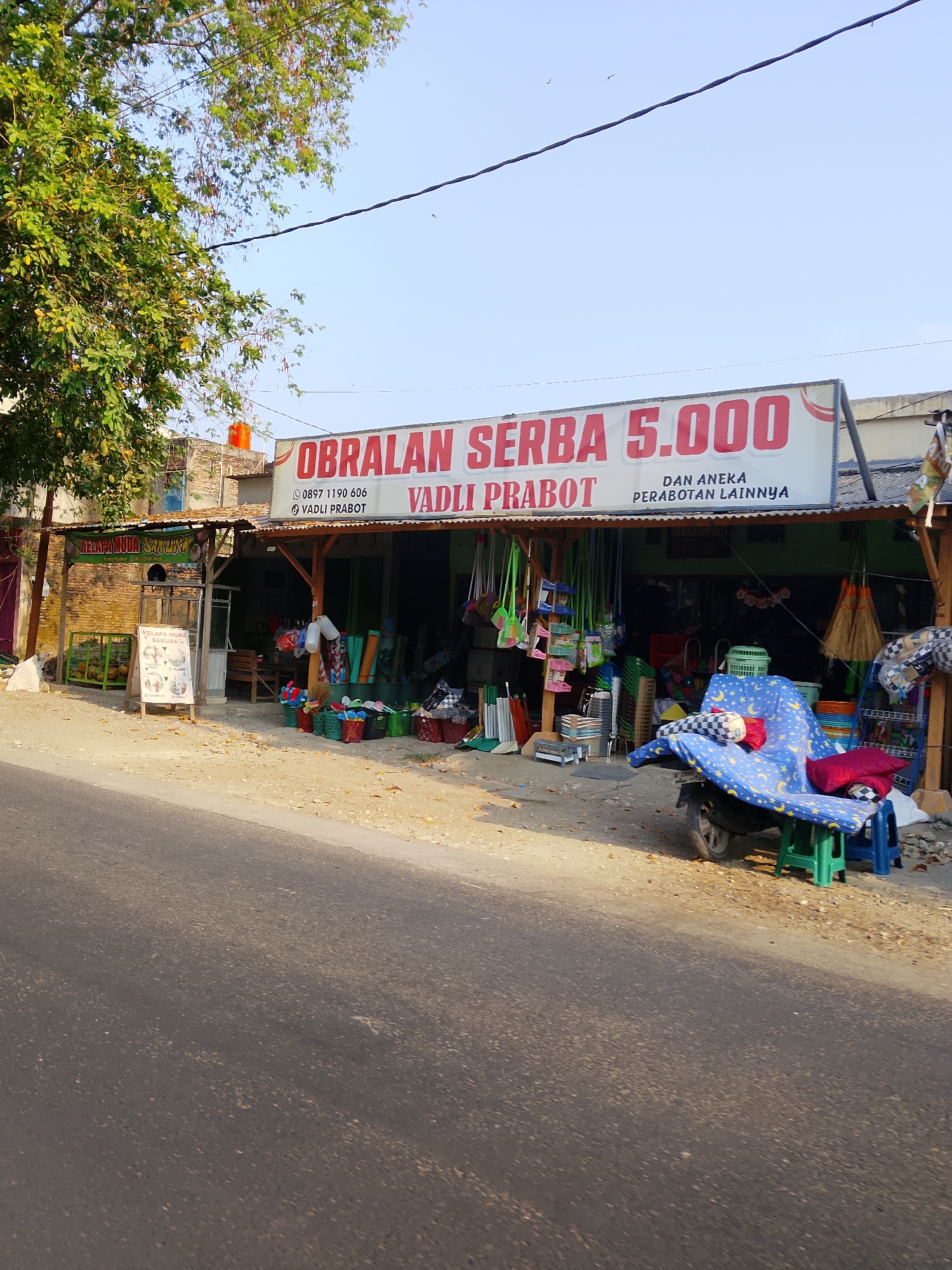 The image depicts a street-side shop in Kasiman, Indonesia, specializing in household goods, as indicated by the sign that reads "OBRALAN SERBA 5.000" (Sales of Everything for 5,000) and "VADLI PRABOT" (Vadli Housewares) with the phone number 0897 1190 606. The shop is open-fronted with a corrugated metal roof supported by wooden beams. Various items are displayed outside, including brooms, mops, buckets, baskets, and other domestic items. A motorcycle is parked in front of the shop, partially covered with a blue and yellow star-patterned cloth, and adorned with cushions. To the left of the shop, there's a sign advertising "KELAPA MUDA SAKURA" (Young Coconut Sakura) and a display of coconuts. The street in the foreground is asphalt, and the ground next to the shop is dirt and gravel. A tree with green foliage is visible to the left, and the sky above is clear and blue. There are no people clearly visible in the image. The scene appears to be set during daylight hours, possibly late afternoon given the angle of the sunlight casting shadows.