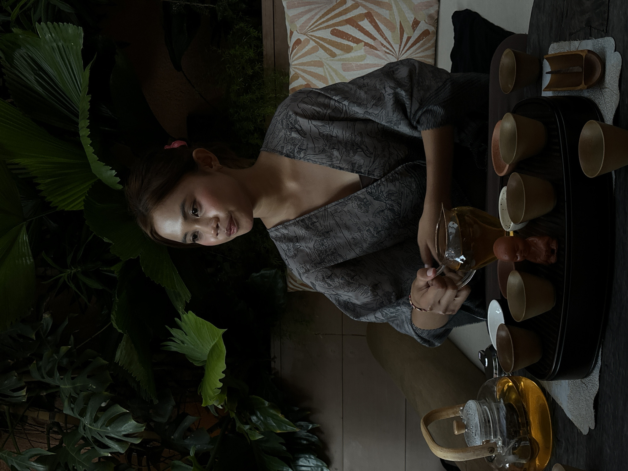 A young woman with long brown hair and fair skin, wearing a grey patterned kimono-style top, pours tea from a glass pitcher into a small cup. She is positioned beside a dark wooden table containing a black tray with several small tea cups and a wooden implement for the tea ceremony. The background includes lush greenery, suggesting a tropical or garden setting. A patterned cushion is visible behind the woman. The scene takes place outdoors, possibly in Pecatu, Indonesia.