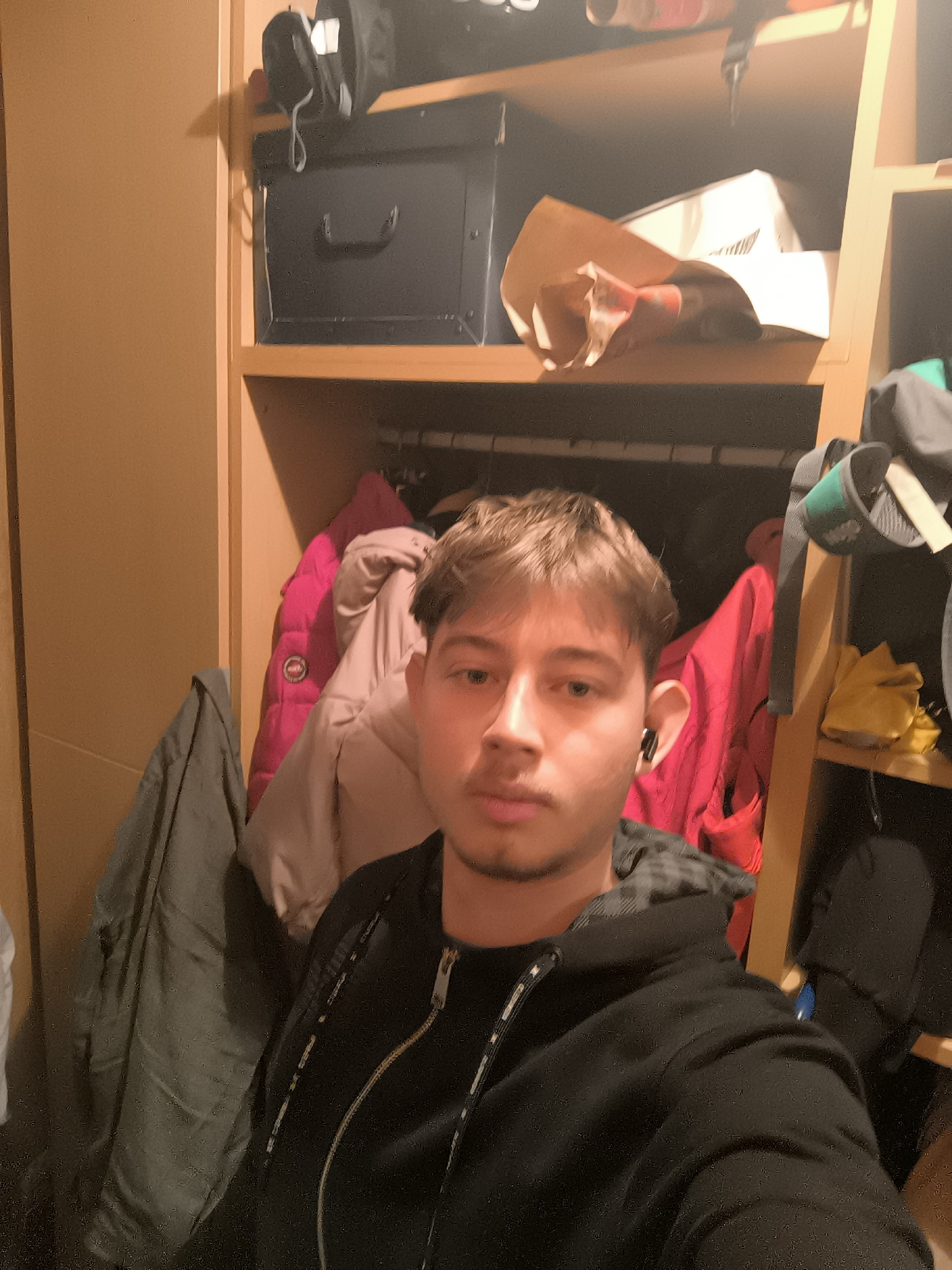 A young man, likely in his late teens or early twenties, is captured in a selfie in an indoor setting, which appears to be a closet or storage area. He has light brown hair, a neutral expression, and light facial hair. A dark earbud is visible in his right ear. He is wearing a black, zipped-up hooded sweatshirt.
The background features a light brown wooden shelving unit or closet. On the top shelf to the left, a black bag and a dark, rectangular box with a handle are stored. To the right on the same shelf, a brown paper bag and a white box are visible. Below these shelves, various articles of clothing are hanging. These include a dark grey jacket on the far left, and a prominent bright pink puffer jacket with a small, unreadable round logo (resembling Canada Goose) in the center, alongside other lighter-colored garments and a red jacket. On a lower shelf to the right, a pair of yellow gloves and a black mitten are stored, with a hint of a blue object beneath.
The lighting is artificial and somewhat dim, casting shadows that suggest an interior space rather than natural daylight. The man is simply taking a photograph, with no other discernible activity. No specific emotional expression is evident. Although the context provided indicates the location is Sofia, Bulgaria, there are no visual cues within the image to confirm this. No clear, legible text is visible in the frame.