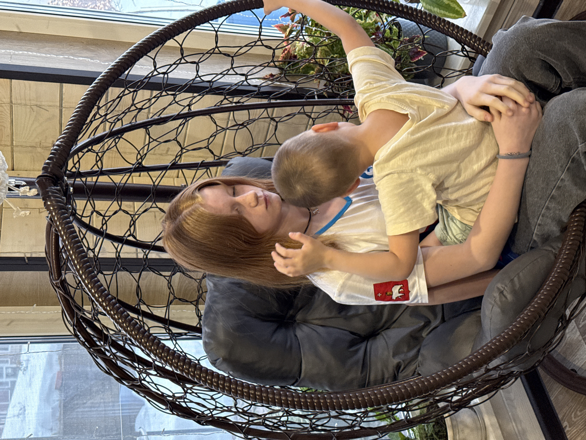 In an intimate indoor scene, two children are nestled together in a large, dark brown hanging chair with intricate woven patterns and soft grey cushions. An older child, likely a girl with long, light brown hair, is lying back with her eyes closed, appearing to be resting or asleep. She wears a white t-shirt with a blue trim around the neckline and a dark necklace.
A younger child, a boy with short, light brown hair, is leaning over her, his head near her neck, suggesting a comforting or affectionate gesture. His left arm is wrapped around her, and his right hand is gently holding hers. He is dressed in a light yellow t-shirt, which has a distinctive red rectangular patch on the right side depicting a white stylized animal head, possibly a moose or reindeer, along with a small white cross. He also wears dark grey pants.
The chair is positioned near a large window, allowing natural light to illuminate the scene, suggesting it is daytime. Outside the window, blurred architectural elements and some light-colored structures are visible, possibly indicating an urban or suburban setting. Lush green and red-leafed plants are also visible in the background, adding a touch of nature to the indoor space. The overall atmosphere is one of warmth, comfort, and sibling affection. The location context for this image is Chaikovsky, Russia.