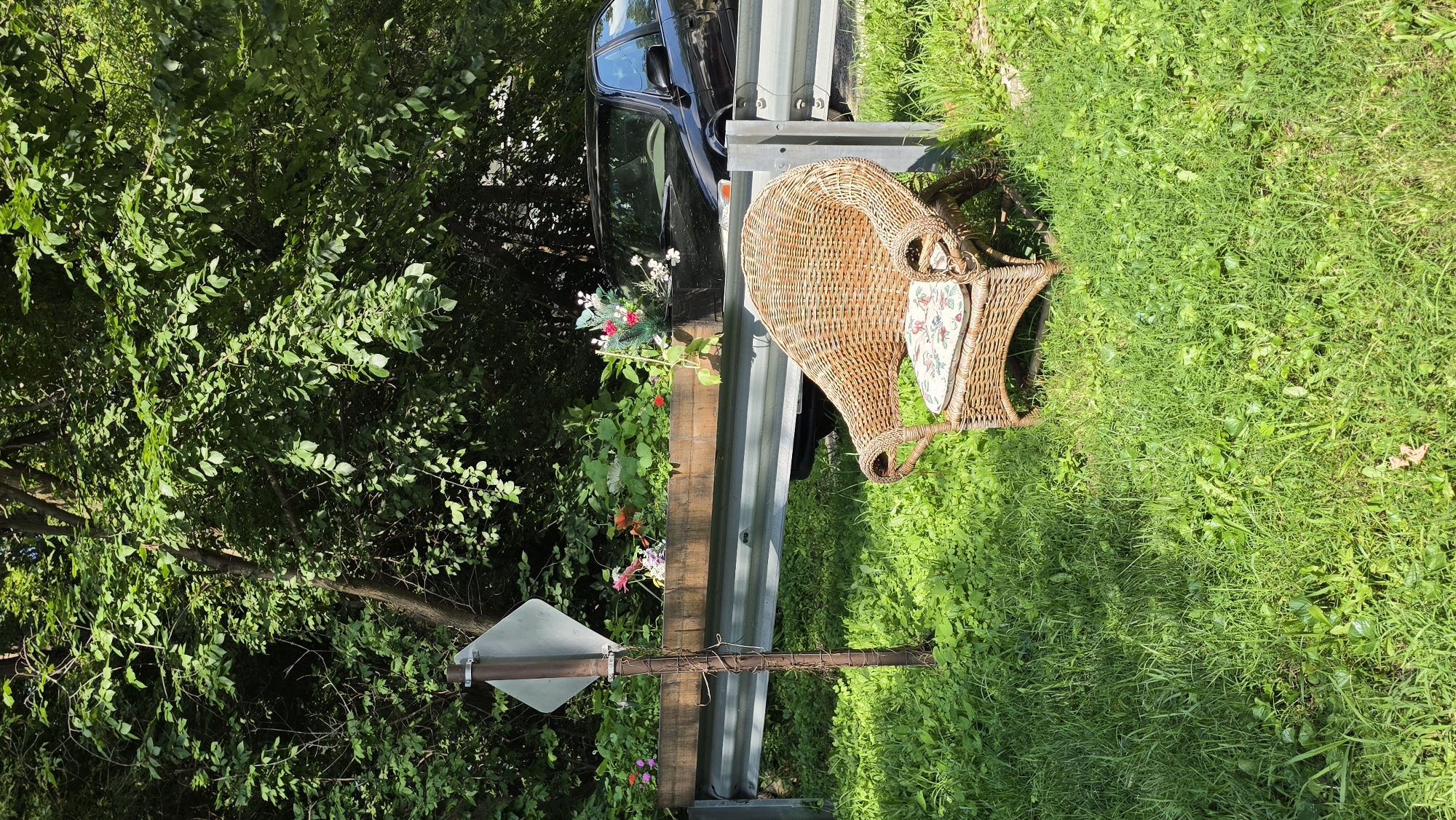 The image depicts an outdoor scene in Elgin, United States of America, featuring a wicker armchair placed on lush green grass. Behind the chair, a wooden planter box filled with various flowers sits atop a metal guardrail. A reflective diamond-shaped signpost is partially visible on the left, next to the planter. A dark-colored car is parked behind the guardrail. The background is dominated by dense green foliage and trees, suggesting a natural or park-like setting. The lighting indicates it is daytime, with sunlight casting shadows on the grass. There are no people visible in the scene.