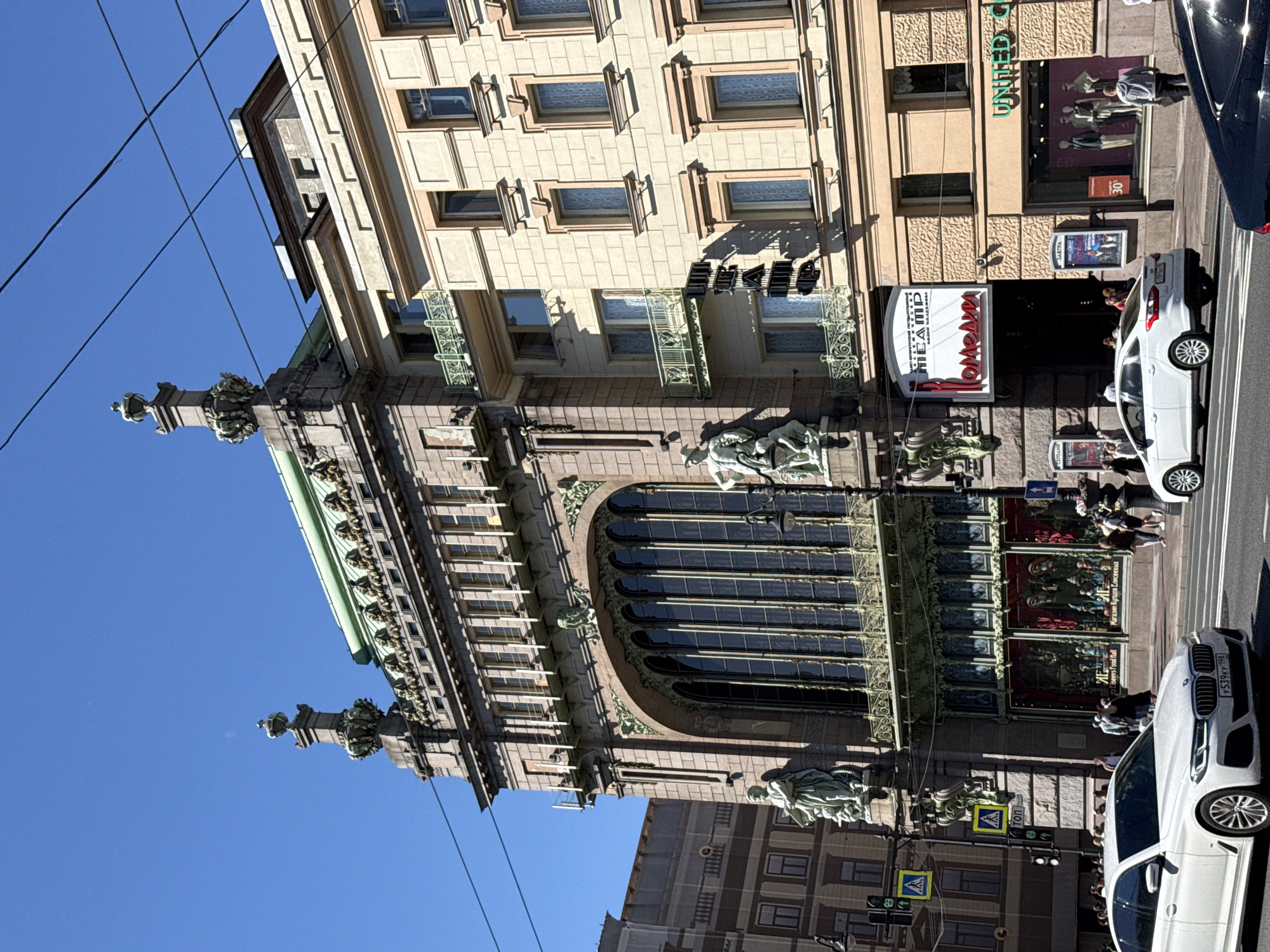 This image captures a sunny day in Saint Petersburg, Russia, focusing on the Singer House, also known as Dom Knigi (House of Books), a prominent Art Nouveau building located on Nevsky Prospekt. The ornate facade of the Singer House is visible, featuring large arched windows, decorative sculptures, and intricate stonework. To the right, a lighter-colored building stands adjacent to it, with multiple windows and balconies.
In the foreground, a white BMW sedan drives on the street, its headlights on. Several other vehicles and pedestrians are present on the street and sidewalks. Traffic lights and street signs are visible, indicating an urban setting. The sky is clear blue. The overall scene depicts a bustling city street with a focus on the architectural grandeur of the Singer House.