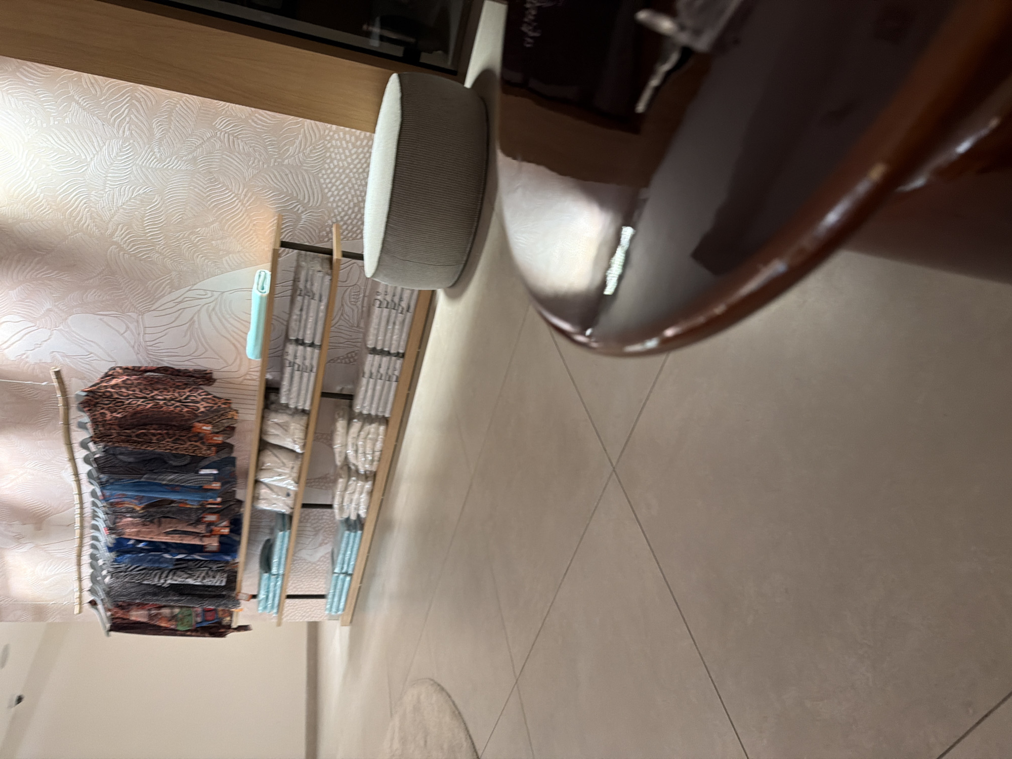 The image depicts the interior of a retail store. In the background, clothing is displayed on a wall-mounted rack, with various patterned shirts hanging. Below the clothing rack, shelves are stocked with folded garments, appearing to be folded stacks of towels or similar textiles. To the right, a round, light-colored ottoman sits on the tiled floor. In the foreground, a glossy, dark brown surface, possibly a tabletop, reflects light and appears to have some items on it, one of which resembles a key. The overall environment is clean and well-lit, suggesting a commercial space. No specific details about Boreti, Montenegro, are evident in the image itself.