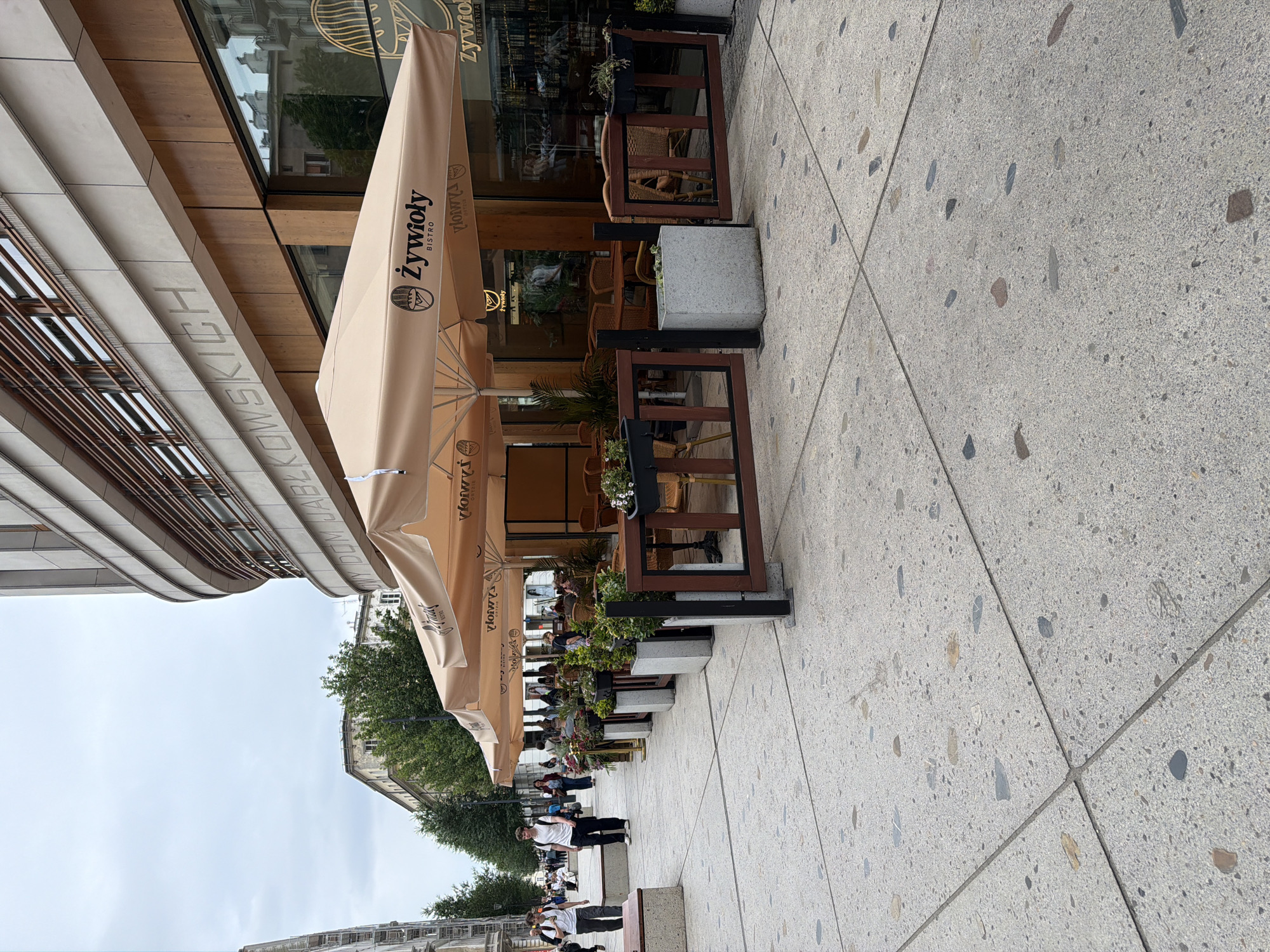 An outdoor seating area of a bistro, identified by the name "Żywioly Bistro" visible on the beige umbrellas, is set up on a wide, gray, speckled sidewalk. The bistro has a modern facade with large windows and wooden accents. Several tables and chairs made of wicker and wood are arranged under the umbrellas, with potted plants and wooden barriers separating the seating area from the sidewalk. In the background, pedestrians are walking along the sidewalk, and there is a building with a curved facade and horizontal wooden slat detailing. The sky is overcast.
