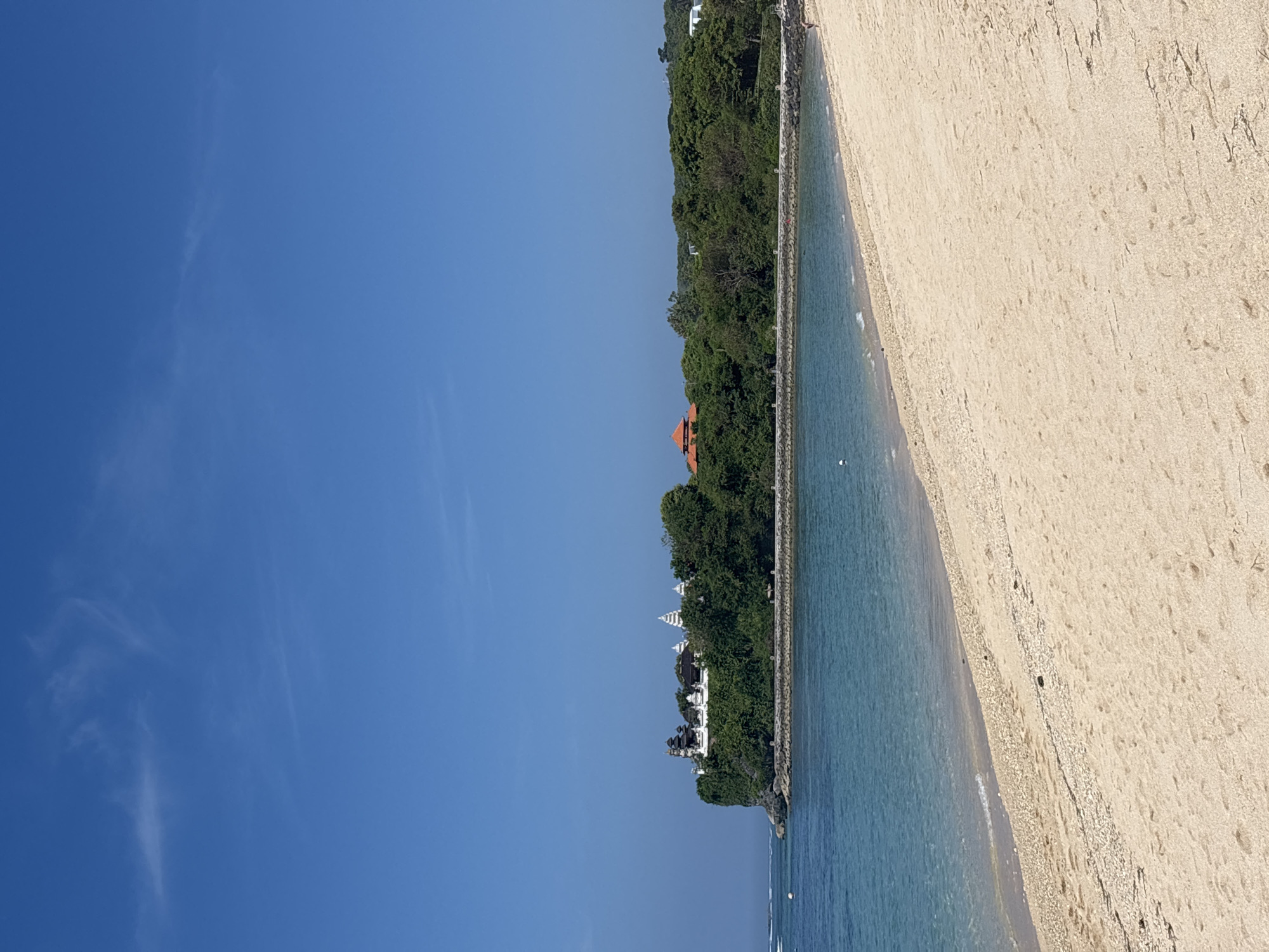 The image displays a vibrant coastal landscape, initially presented rotated 90 degrees clockwise. When viewed correctly, the scene features a clear blue sky, a tranquil ocean, a sandy beach, and a lush, tree-covered coastline dotted with distinctive architecture, characteristic of Kuta Selatan, Indonesia.
The upper section of the image is dominated by an expansive, brilliant blue sky, showing only a few faint, wispy white clouds, indicating excellent, clear weather conditions. Below the sky, the calm ocean stretches across the mid-ground, exhibiting translucent shades of light blue and turquoise near the shore, suggesting clear waters. A prominent stone seawall or breakwater runs horizontally, forming a clear demarcation between the water and the sandy beach. Gentle, small waves are visible breaking softly against this barrier. Further out in the water, a few small, white buoys or floats can be observed.
To the left of the water, a verdant hill or cliff rises, densely covered with tropical trees and foliage. Several structures are visible nestled within this greenery. Most notably, there are multiple white buildings with distinct multi-tiered roofs, strongly resembling Balinese temples (Pura), which serve as a significant cultural and architectural landmark. Further along the coastline, a building with a distinctive orange-red tiled roof can also be seen through the trees.
The right side of the image is occupied by a wide expanse of light-colored sandy beach. The sand appears clean but shows subtle patterns of footprints and ripples, indicating it has been recently traversed. The overall impression is one of a peaceful, beautiful tropical setting on a bright, sunny day, evoking a sense of calm and natural splendor. There is no visible text in the image.