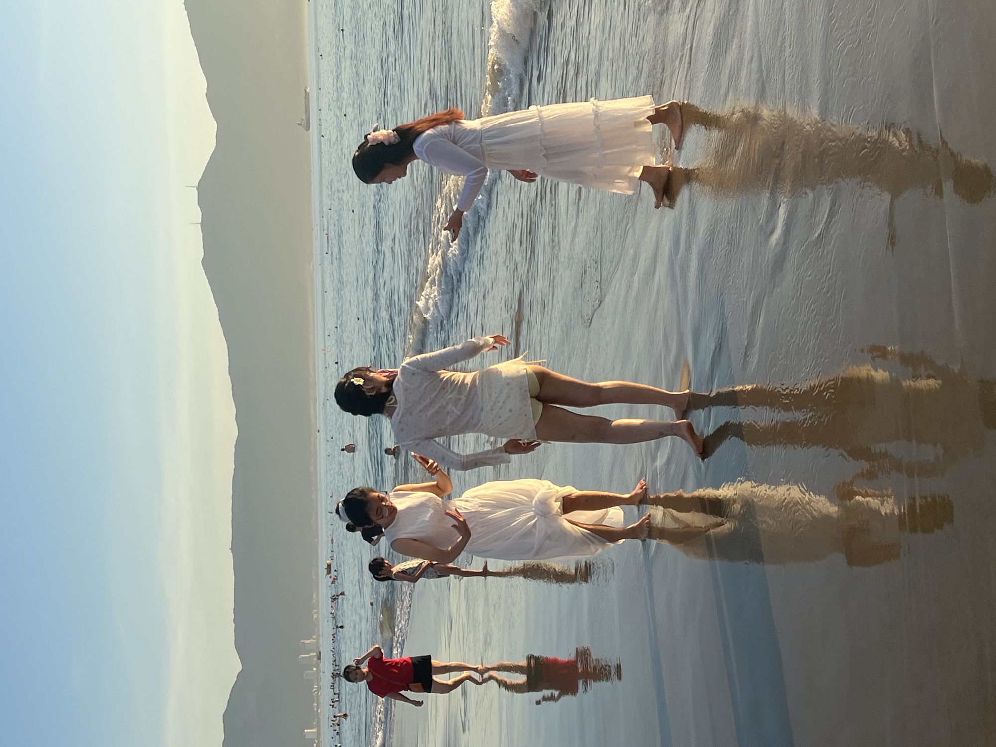 The image captures a serene sunset scene at My Khe Beach in Da Nang, Vietnam. The golden light illuminates the wet sand, creating reflections of the people present. In the foreground, three young women are visible. One woman on the left wears a white dress and is looking down at her hands. To her right, another woman in a white, sheer dress and light-colored shorts is interacting with the first woman. On the far right, a third woman, also in a white dress, is facing the other two, with her hand outstretched. In the background, several other individuals are scattered along the beach and in the shallow water, with a hazy mountain range forming the backdrop. The ocean is calm, with gentle waves lapping at the shore. The sky is a soft blue, transitioning into a lighter hue near the horizon.