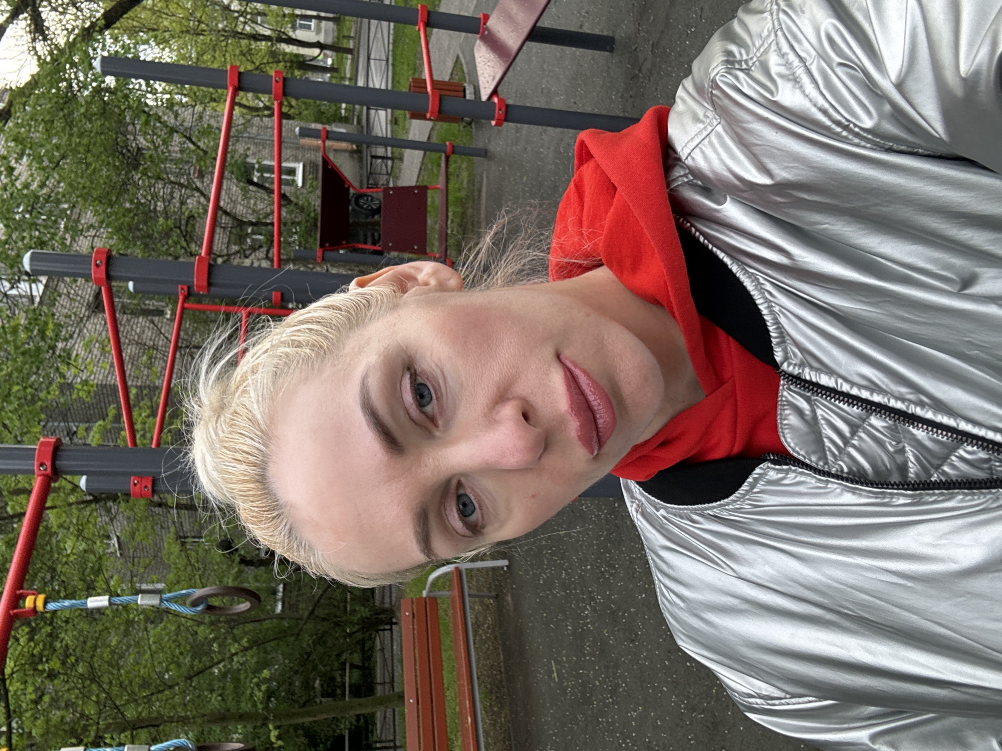 A woman with blonde hair stands in front of a playground. The playground has a climbing frame with red and grey bars and swinging rings suspended from blue ropes. Behind the climbing frame are green trees and a building. To the side is a brown wooden bench. The woman is wearing a red hoodie and a shiny silver jacket.