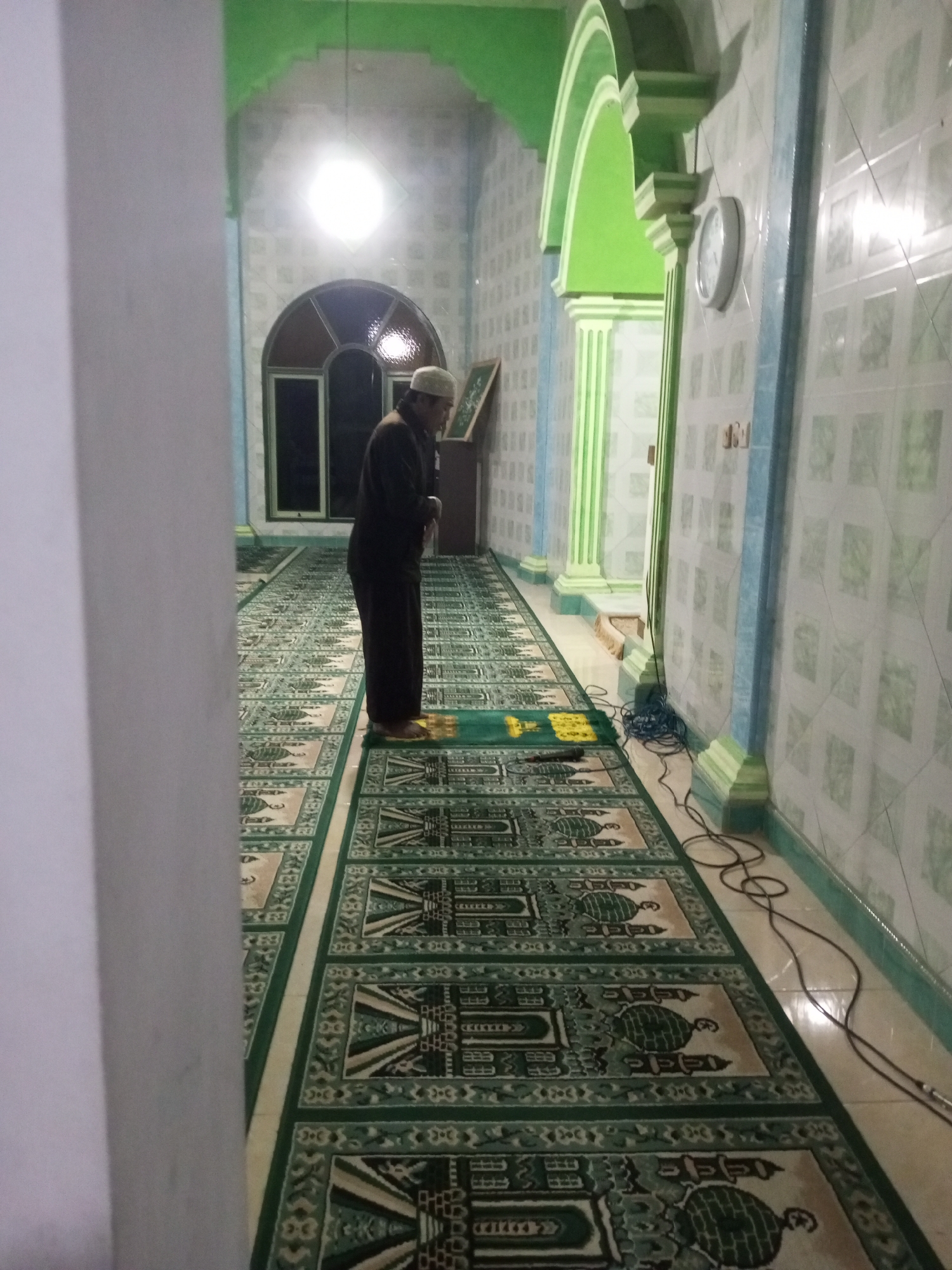 A man wearing a white skullcap and a dark thobe is standing on a prayer rug in the hallway of a mosque. The hallway is decorated with green and white patterned tiles on the walls and a patterned carpet on the floor. Arched doorways and pillars with green accents line the hallway. A clock is mounted on the wall. Electrical cords are visible on the floor. The scene is lit by artificial light. The location is the mosque in Andongsili, Indonesia.