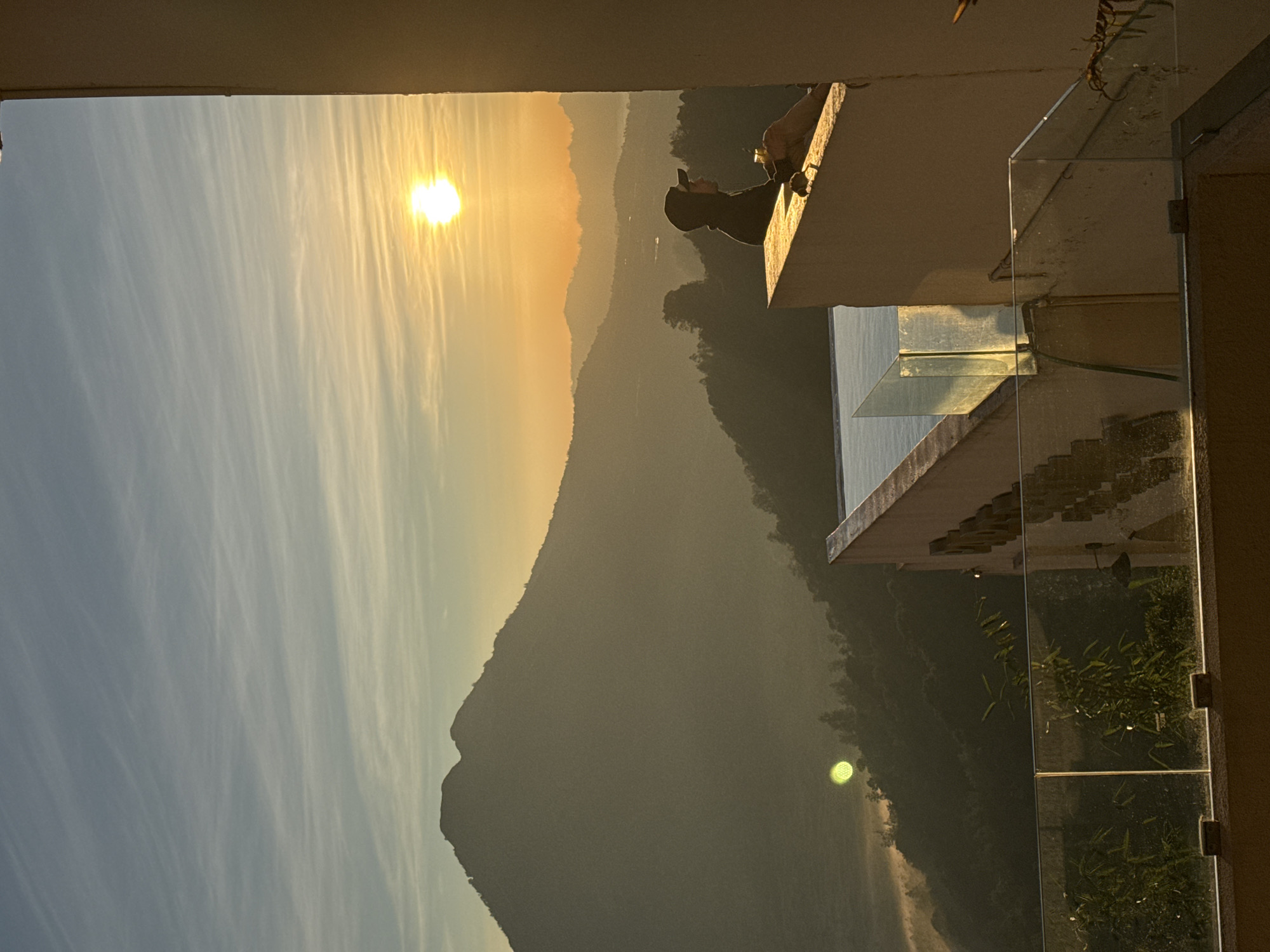 This vertical image displays a mountainous landscape in Kintamani, Indonesia, during sunrise. A prominent, dark silhouette of a mountain, consistent with Mount Batur, dominates the left side of the frame. The sky transitions from pale blue with wispy cirrus clouds at the top to a bright golden-orange along the horizon, where the radiant sun is positioned on the upper right. Distant mountains and valleys are layered and partially obscured by atmospheric mist, indicating an early morning setting.
In the foreground-right, a person is seated on a light-colored architectural parapet, facing towards the left. The individual is wearing a dark hooded top and a baseball cap, holding a clear glass in their right hand. A segment of an infinity pool is visible behind the parapet, reflecting the sky. Below the parapet, a glass safety railing extends across the frame. A distinct green circular lens flare is present in the lower-left portion of the image, within the misty valley. A large, light-colored building wall frames the right side of the image.