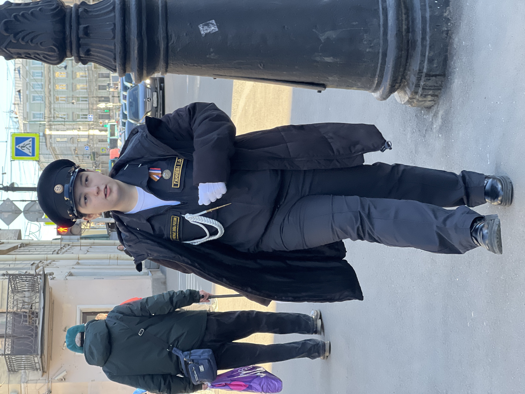 A young person in a dark naval uniform with a black cap stands beside a black lamppost in Saint Petersburg, Russia. The uniform includes name tags in Cyrillic script. A person in a dark green coat walks by in the foreground. Buildings, vehicles, traffic lights, and a pedestrian crossing sign are visible in the background.