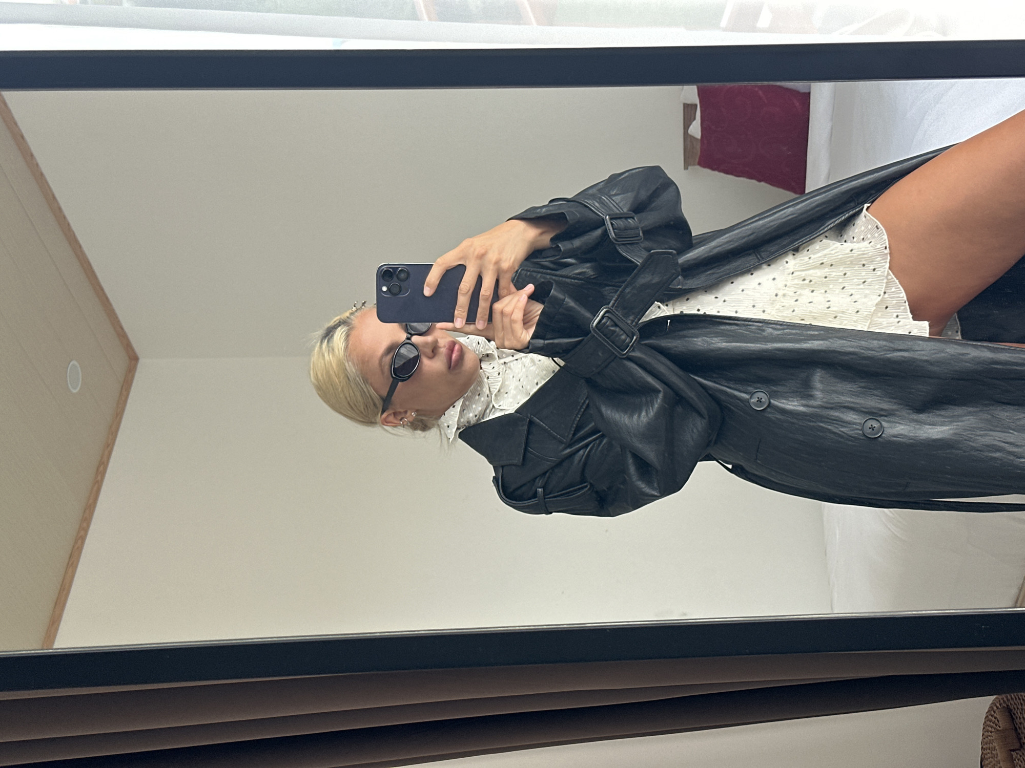 A young woman with blonde hair tied back is taking a selfie in a large mirror. She is wearing large oval sunglasses, a white ruffled shirt with black polka dots, and a long black leather trench coat. She is holding a dark-colored smartphone in her right hand to take the photo. The background of the reflection shows a bedroom with a white bed, a red pillow, and a wooden headboard. There is a tall, dark-framed mirror in the foreground, and part of a brown curtain is visible to the left of the mirror. The woman's location is Kuta Selatan, Indonesia, likely within a hotel room or apartment given the bedroom setting.