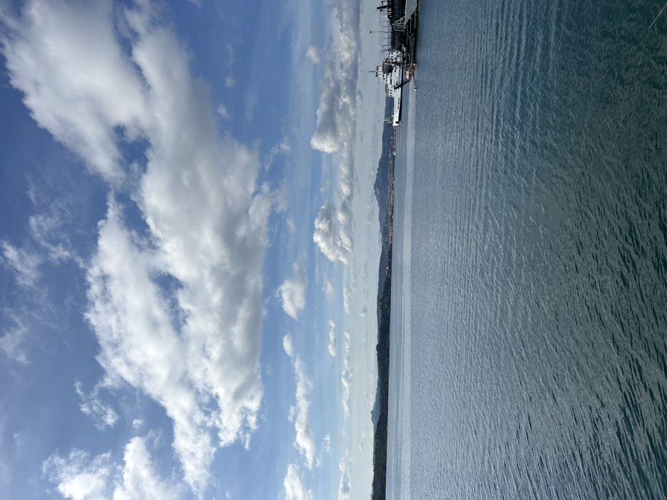 This image captures a wide maritime scene near the city of La Maddalena, Italy, specifically from the vicinity of the La Maddalena archipelago, likely looking towards the Strait of Bonifacio and the northern coast of Sardinia.
The upper half of the frame is dominated by a bright blue sky partially covered by numerous scattered white cumulus clouds of varying sizes and densities. Some higher, thinner cirrus clouds are also visible. The lower half features a vast expanse of dark blue-green water with subtle ripples across its surface, indicating calm conditions.
Along the horizon, a series of dark, low-lying landmasses, consistent with islands or coastal terrain, are visible. To the left, a vegetated island appears relatively close. Further in the mid-ground and extending to the right, a more distant, mountainous coastline is present, displaying areas of urban development, likely the city of Palau on the Sardinian mainland.
In the right mid-ground, a white and blue passenger and vehicle ferry, bearing the name "DELCOMAR" on its side, is prominently docked or moored. Adjacent to it are several smaller, darker-hulled vessels, possibly service boats or other harbor craft. No individuals are discernible on any of the vessels or along the distant shore. The scene is illuminated by natural daylight, suggesting clear weather.