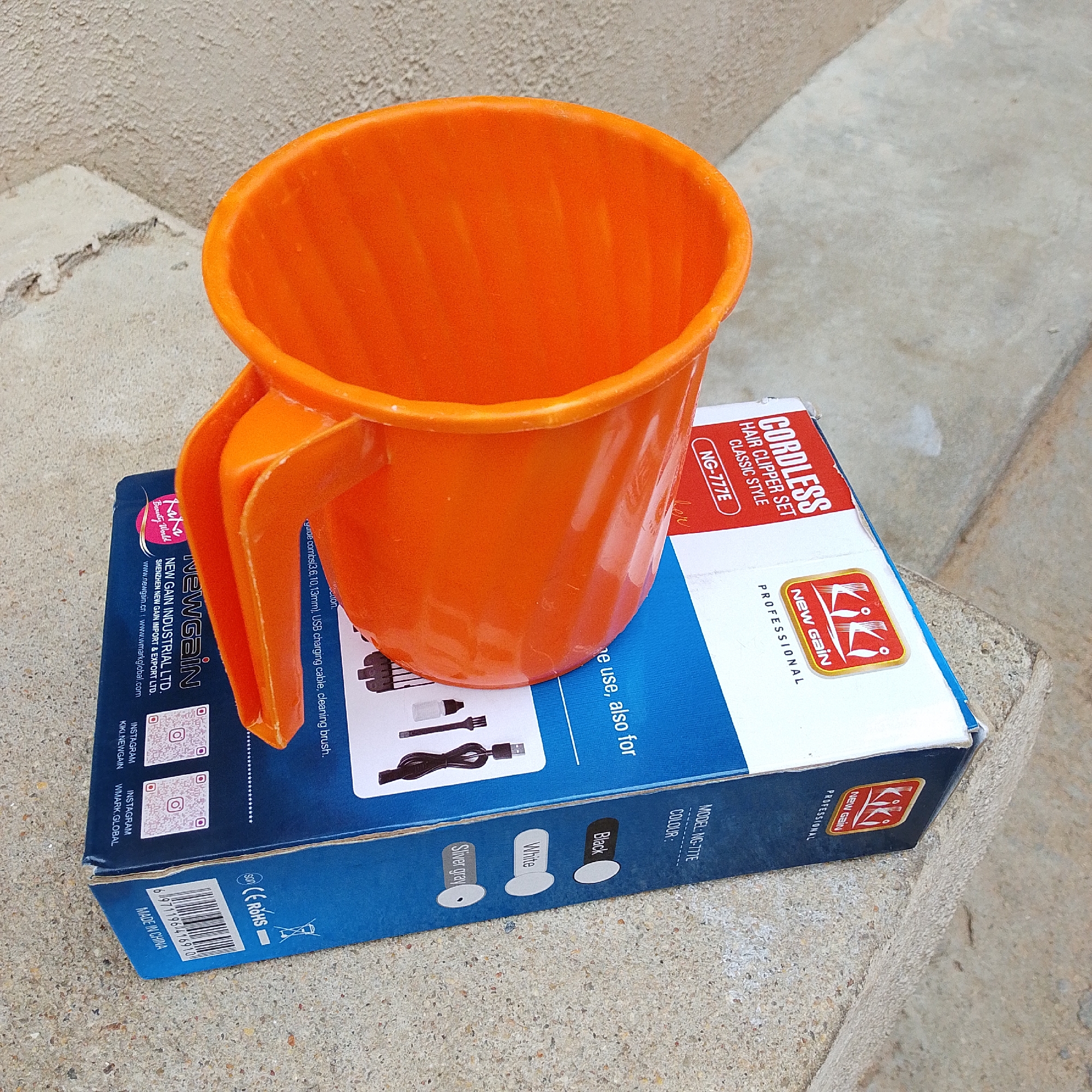An orange plastic mug sits atop a blue and white cardboard box. The box displays text indicating it contains a "CORDLESS HAIR CLIPPER SET CLASSIC STYLE" with the model number "NG-777E". Brand logos and text such as "NEW GAIN INDUSTRIAL LTD.", "NEW GAIN", and "Kiki PROFESSIONAL" are visible on the box. There are also images of various hair clipper accessories and QR codes for Instagram. The mug has a handle and appears to be empty. The scene is outdoors, with a concrete textured surface visible around the objects. The lighting suggests it might be daytime. The location context is Jalingo, Nigeria.