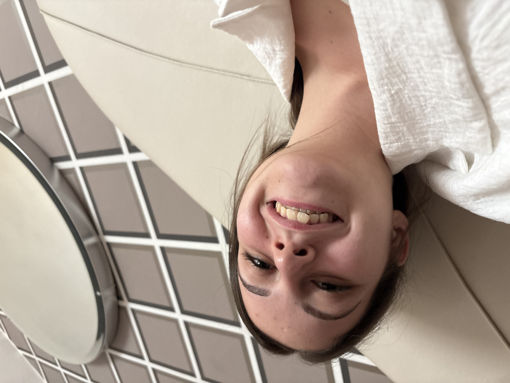 An upside-down portrait captures a young female smiling broadly at the camera. She has dark hair and is partially wrapped in a white, textured towel or robe around her head and shoulders. Braces are visible on her upper teeth, contributing to her cheerful expression. She appears to be lying on a light beige, upholstered surface, possibly a bed or sofa. The background features a distinctive tiled wall with a geometric pattern of light gray and darker gray squares and rectangles, accentuated by white grout lines. On the left, a round object with a metallic rim, potentially a mirror or light fixture, is partially visible against the tiled surface. The scene suggests an indoor setting, likely a modern room, possibly a hotel room or bathroom, located in Burgas, Bulgaria. The bright, even lighting implies daytime or well-lit conditions. The subject's wide smile conveys happiness and a playful mood. No text is visible in the image.