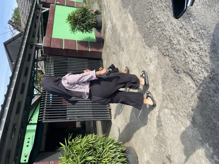 The image shows a woman in a black hijab and a black-and-white patterned long-sleeved shirt walking on a concrete path in front of a house in Jatiluhur, Indonesia (latitude: -6.576, longitude: 107.438). She is wearing black pants and black sandals. A small child is walking behind her, also wearing sandals. The woman is holding a black smartphone in her right hand. The house is painted red and green, with a black metal gate and a green metal awning. There is a large potted plant on the left side of the image and a smaller potted plant on the right side of the image. The ground is covered in rough concrete. The image was taken during the daytime. The image is slightly blurred, but it is clear enough to see the details of the scene.