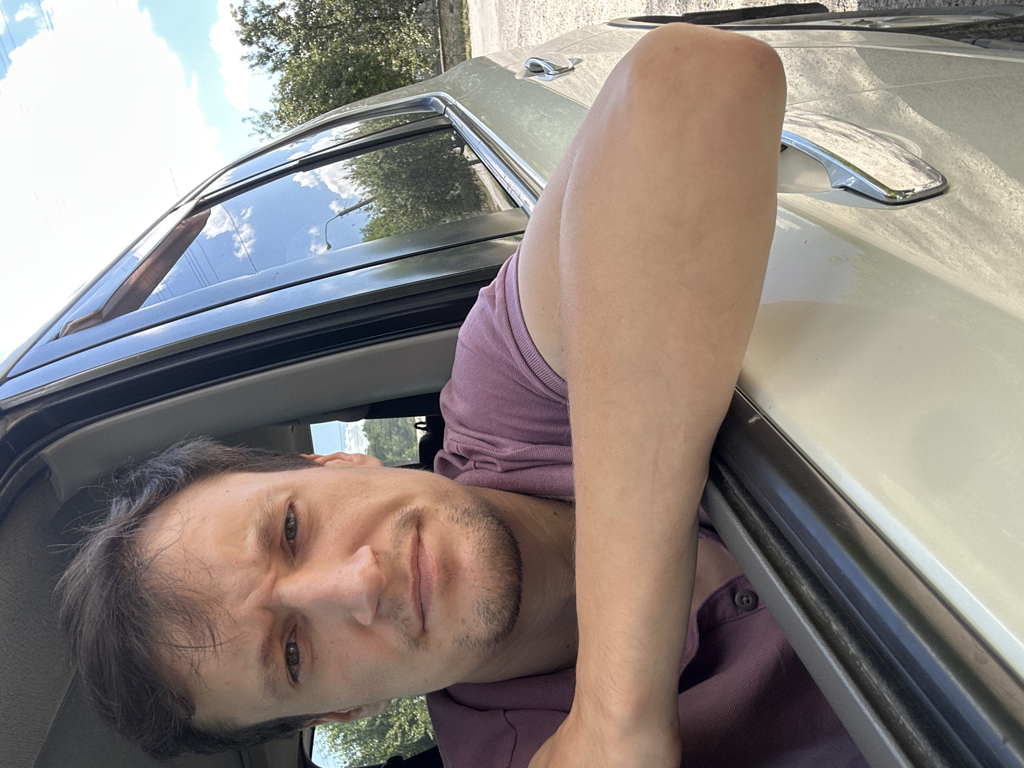 A young man is pictured from the chest up, leaning out of the driver's side window of a car. He has short, dark brown hair and a light beard. He is wearing a purple polo shirt. The car is light-colored and appears to be parked on a dirt or gravel road. Trees and a blue sky with white clouds are visible in the background. The scene is likely outdoors on a sunny day.