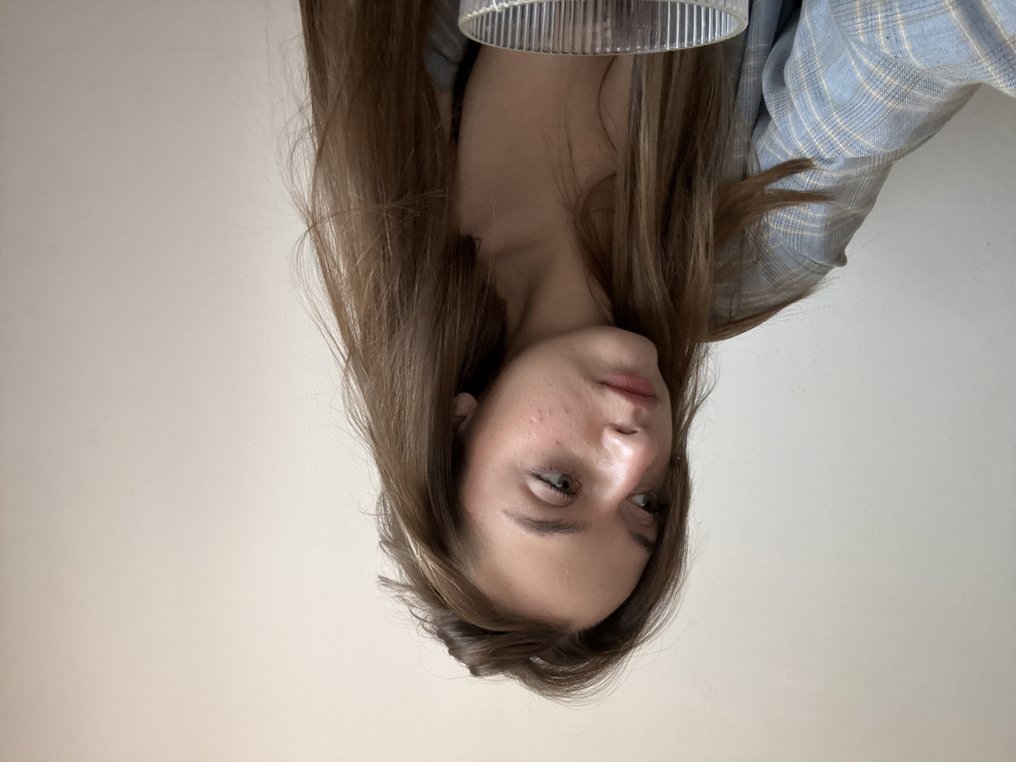 A young woman with light brown hair is pictured from a low angle, tilted. She has fair skin, green eyes, and is wearing a blue plaid shirt. Above her is a clear, fluted glass object. The background is a plain, light-colored wall. The location is not specified, and there is no visible indication the photograph was taken in Kragujevac, Serbia. No actions or interactions are visible.