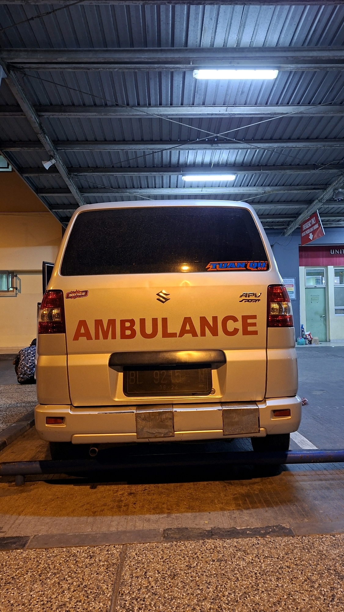 A rear view of a silver Suzuki APV Sport ambulance is captured in a dimly lit covered parking area, likely at a hospital, given the context of an ambulance. The vehicle's license plate is visible and reads "BL 9215 N". Two red reflective stickers are attached to the rear bumper. A sticker on the rear windshield reads "TUAN O.I.B 019". The ambulance is parked on an asphalt surface with speckled paving stones in the foreground. Overhead, a corrugated metal roof is supported by metal beams, with fluorescent lighting illuminating the area. In the background, a building with a red and white sign indicating "EMERGENCY ONLY" and "UNIT" is visible. A person wearing a patterned shirt is sitting on the ground to the left of the ambulance.