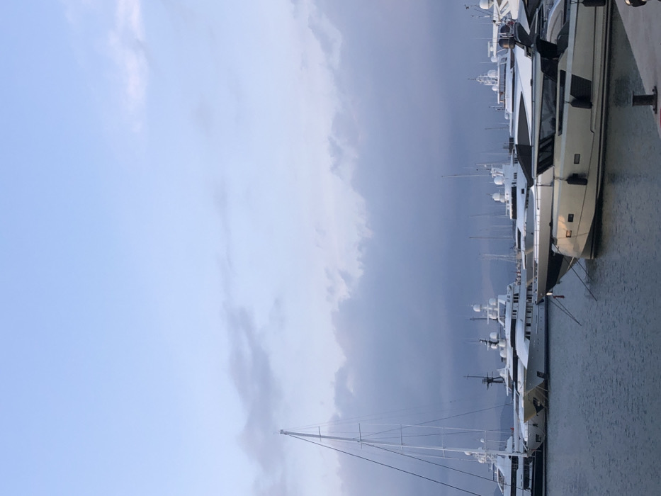 The image shows a marina in Palaio Faliro, Greece (37.929, 23.685). The marina is filled with a line of docked yachts, mostly white in color. The sky is overcast with gray clouds. The water is calm and reflects the gray tones of the sky. The yachts are arranged in a line, with the largest one closest to the camera, its white hull visible. Several other, smaller yachts are further back in the line, partially obscured by the larger boat. There are no people visible in the scene.