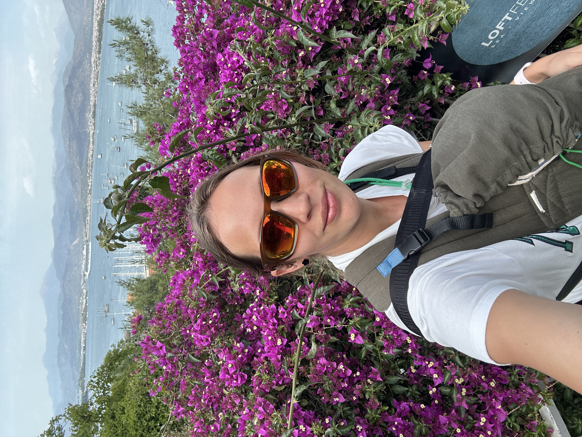 A selfie captures a person wearing sunglasses and a white t-shirt, situated near a vibrant display of purple flowers. The person is wearing what appears to be a baby carrier and an object partially visible in the frame has text that contains the words "LOFT" and "SEA VIEW". In the background, a coastline is visible, with numerous boats dotting the water. The image appears to be taken in Fethiye, Turkey, given the landscape and likely association to LOFT's Fethiye Sea View project.