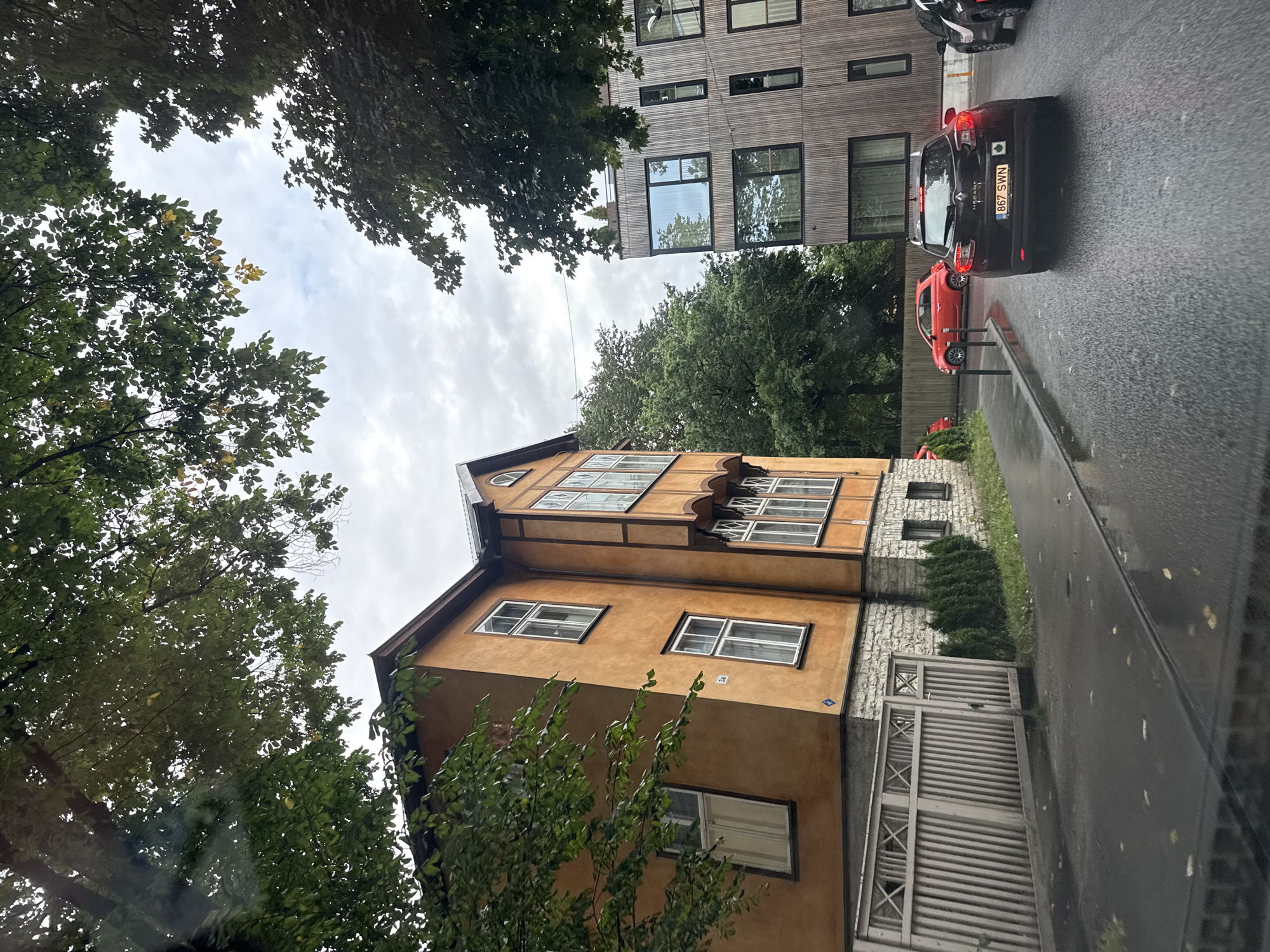The image shows a street scene in Tallinn, Estonia, featuring two buildings and parked cars. On the left is a two-story, orange-colored building with white-framed windows and decorative wooden trim. A white wooden fence runs along the front of this building. To the right of the orange building is a modern, multi-story building with a facade of vertical wooden planks and large, dark-framed windows.
Several cars are visible. A black Renault Megane is in the foreground, facing away from the camera, with the license plate "867 SWN". To its left, a red hatchback is parked. Another dark-colored vehicle is partially visible to the right of the Renault.
The street appears wet, suggesting it may have recently rained. Trees with green leaves overhang the scene, casting shadows. The sky is overcast. The scene is devoid of people.