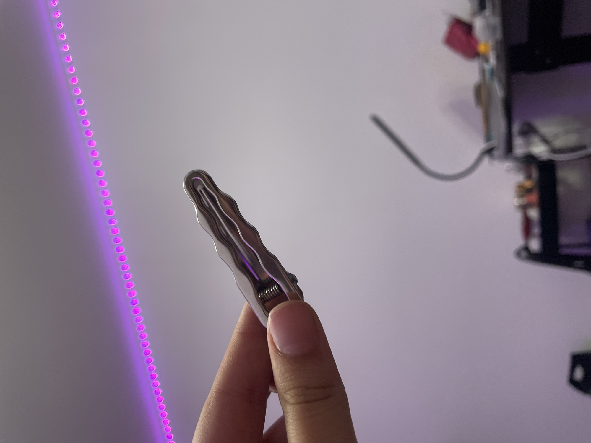 In Kokkorevo, Russia, a person's hand is holding a silver-colored metal clothespin with a coiled spring. The clothespin has a wavy texture. The background features a wall with a string of purple LED lights running horizontally across it. Below the wall, a blurred image of a desk with various objects on it, including what appears to be a lamp and containers, is visible. The lighting in the scene is dim, with the purple LED lights providing the primary illumination.