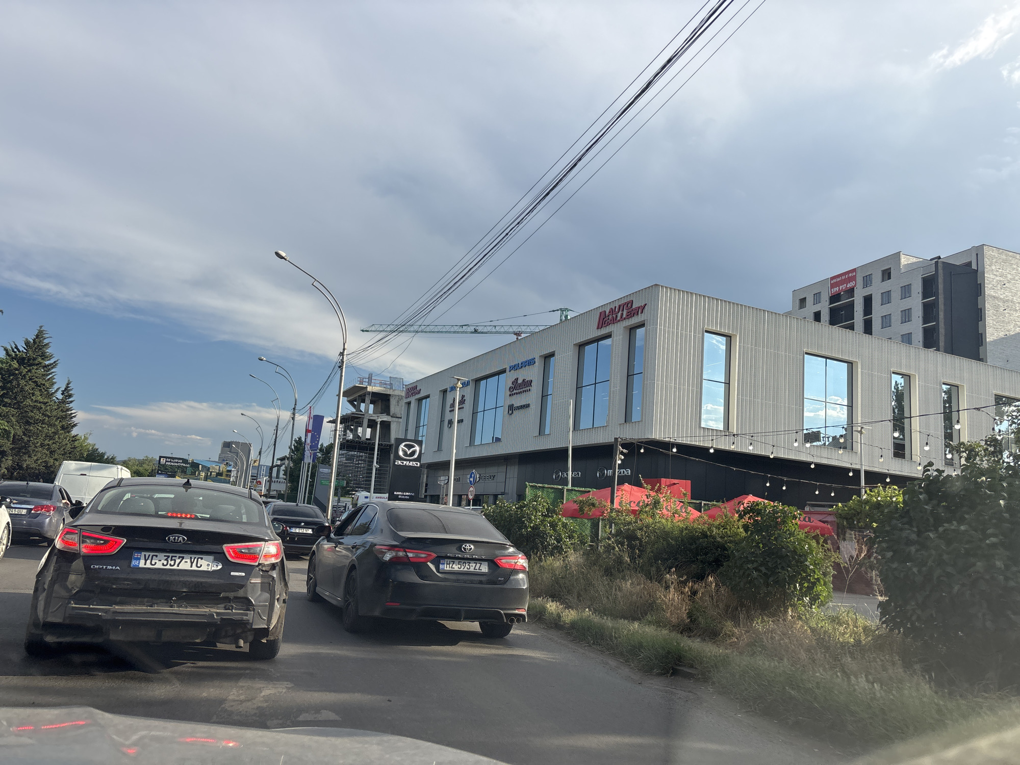 This is a medium-distance, street-level, eye-level, horizontal, color photograph taken from within a vehicle, showing a traffic scene in Tbilisi, Georgia, on a partly cloudy day. The foreground is dominated by the rear of a black Kia Optima with a Georgia license plate reading "YC-357-YC". The car appears to have damage to its rear bumper. To the right of the Kia, a black Toyota Camry with a Georgia license plate "HZ-593-ZZ" is visible. Several other cars are in the traffic queue.
In the background, a modern, multi-story building with a facade of vertical ribbed panels and large reflective windows stands prominently. Signs on the building include "AUTO GALLERY", "POLARIS", and "Indian Motorcycle". A construction crane is visible to the left of the building. Streetlights and overhead power lines crisscross the sky. Red awnings and greenery are situated in front of the building. The scene appears to be on a relatively wide, paved road with traffic.